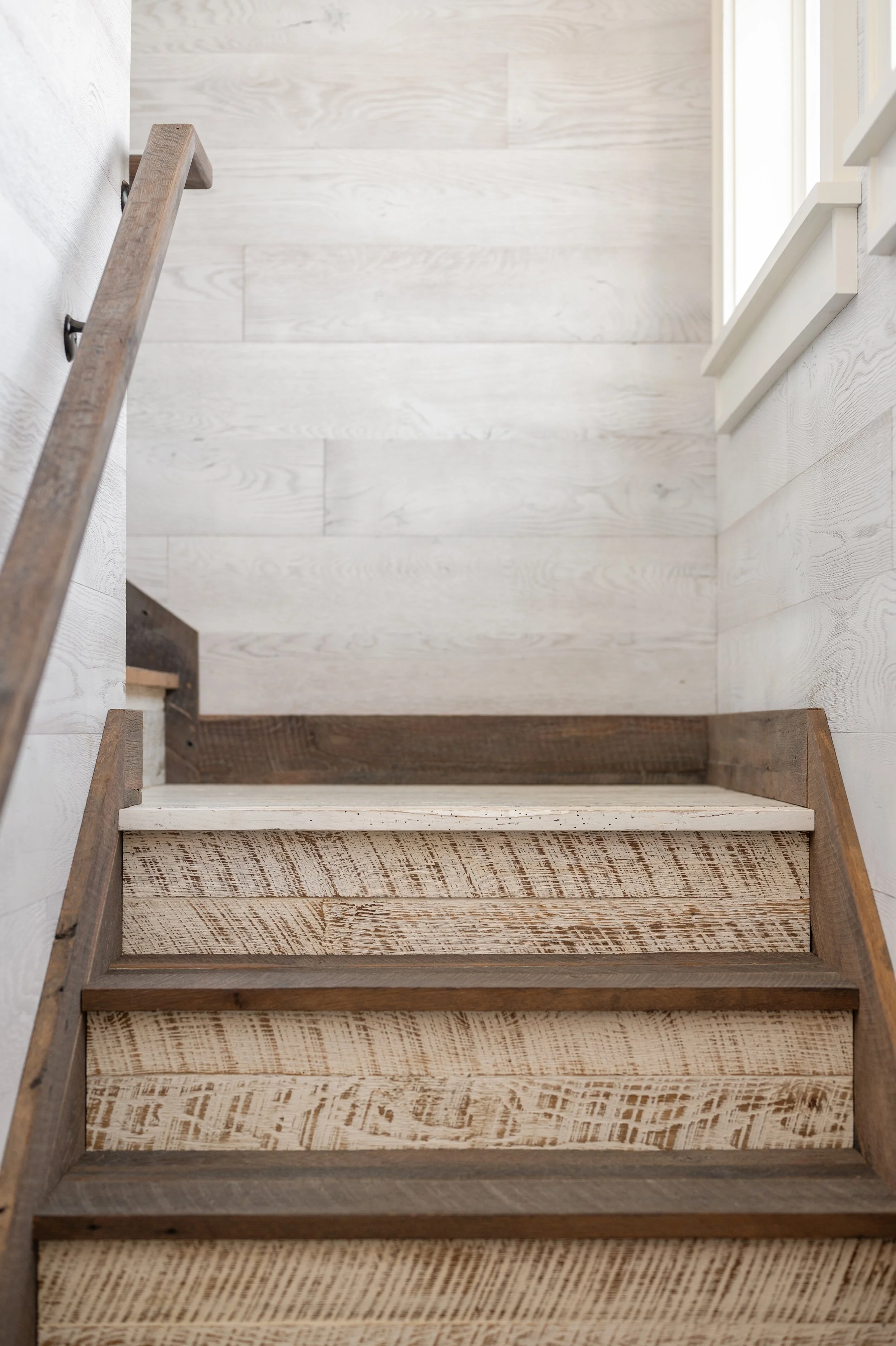 Wooden stairs with a railing leading up to a bright room with white walls and a large window.