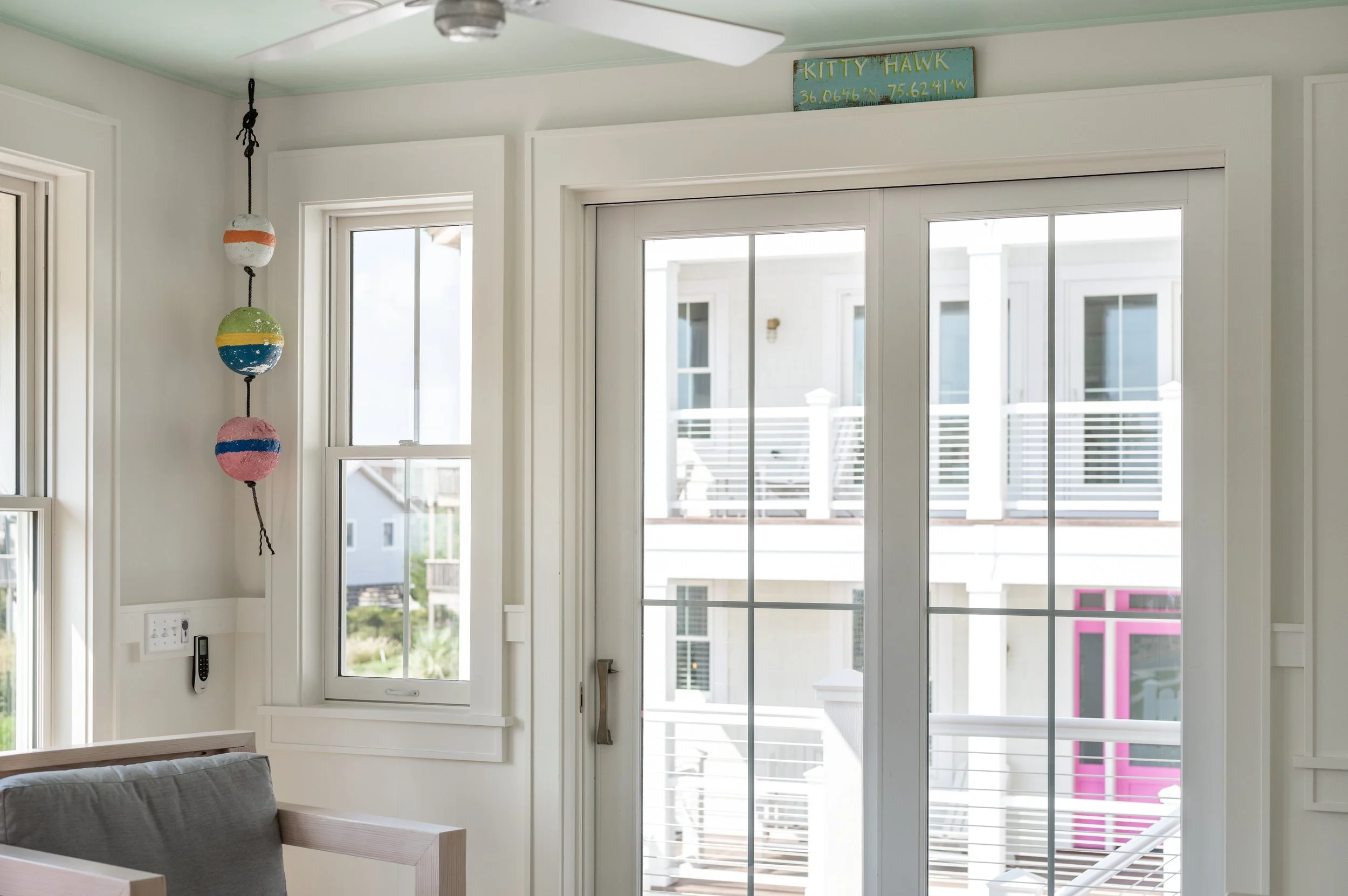 Interior view of a room with large sliding glass doors leading to a balcony or patio, with neighboring white buildings visible outside. The ceiling fan is visible at the top, and a colorful decorative sign reads 'KITTYY HAWK' with coordinates above t