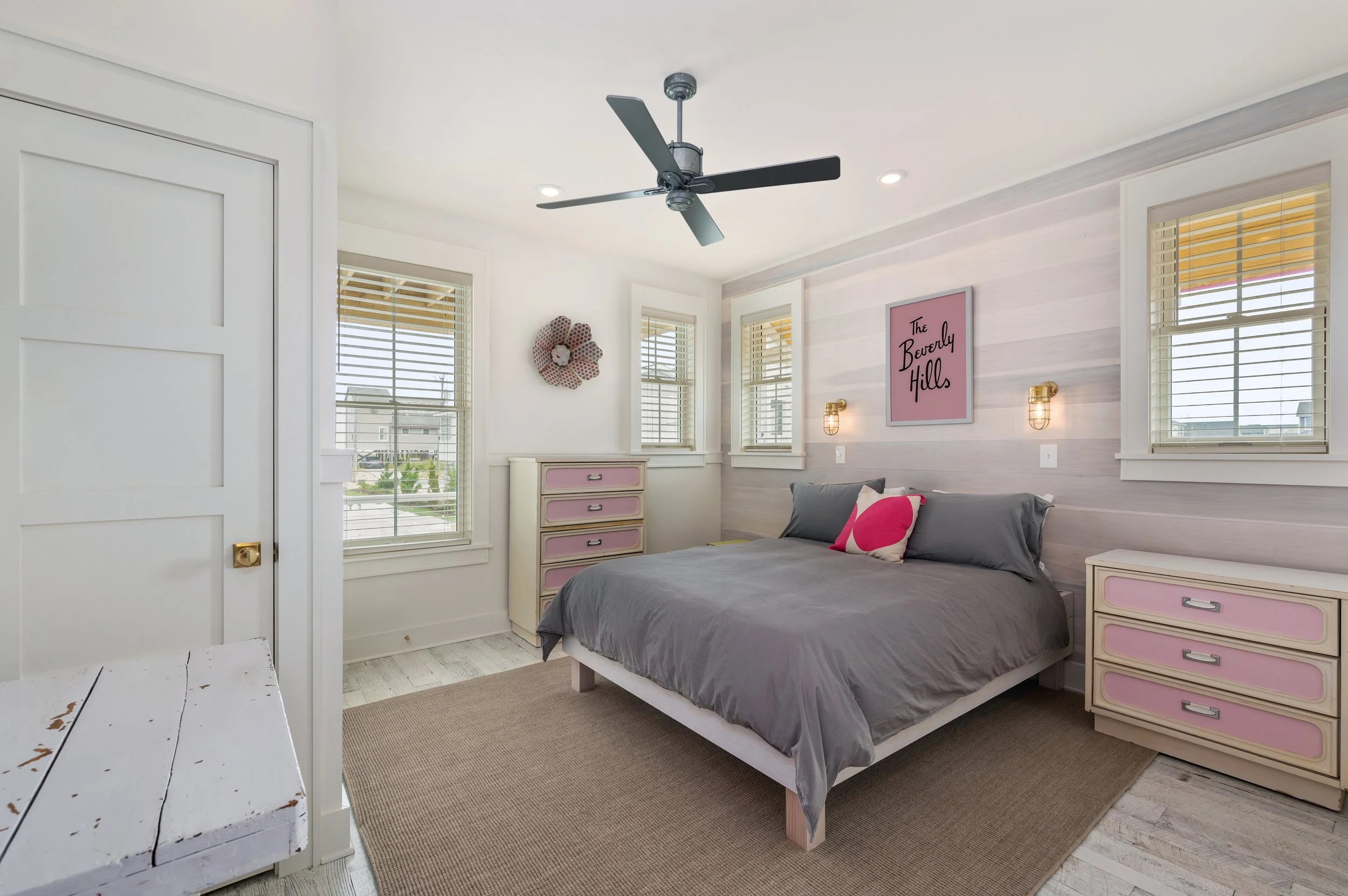 Bedroom with gray bed, pink and gray accent pillows, white furniture with pink accents, three windows with white blinds, wall art with pink background and black text, ceiling fan, beige rug, white wall with light wood paneling.