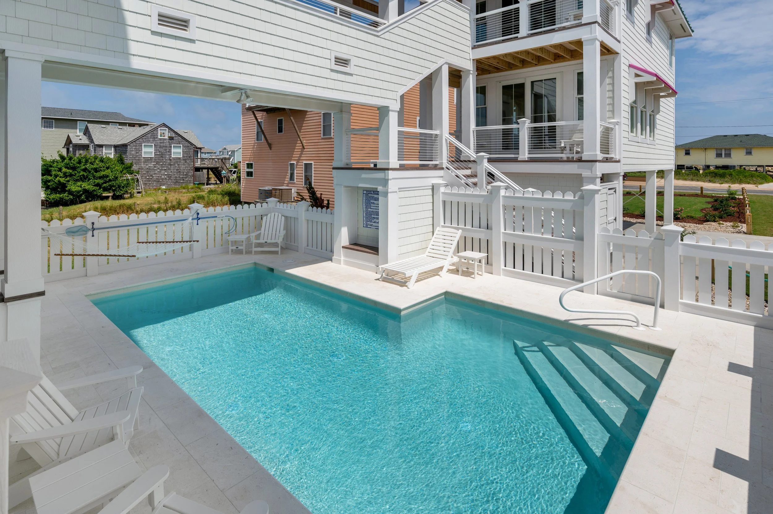 A backyard swimming pool area with white fencing, lounge chairs, and a multi-story house with white siding and balconies, set in a residential neighborhood with other houses visible in the background.