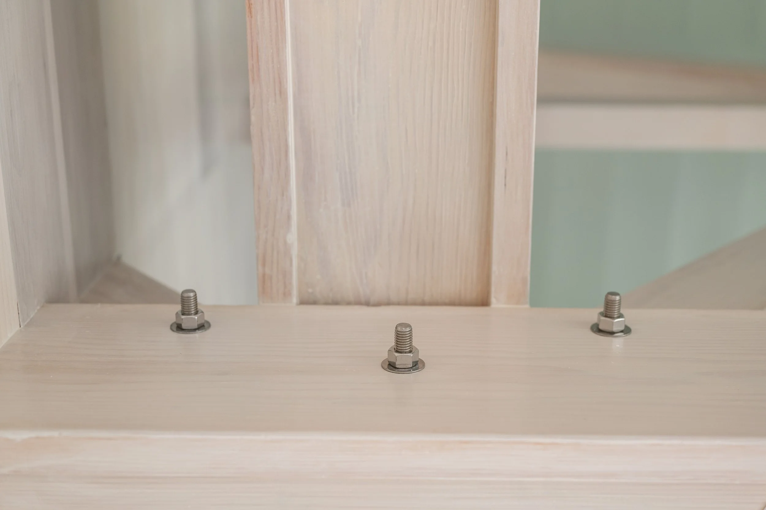 Close-up of three metal bolts on a light wooden surface with a vertical light wood panel in the background.
