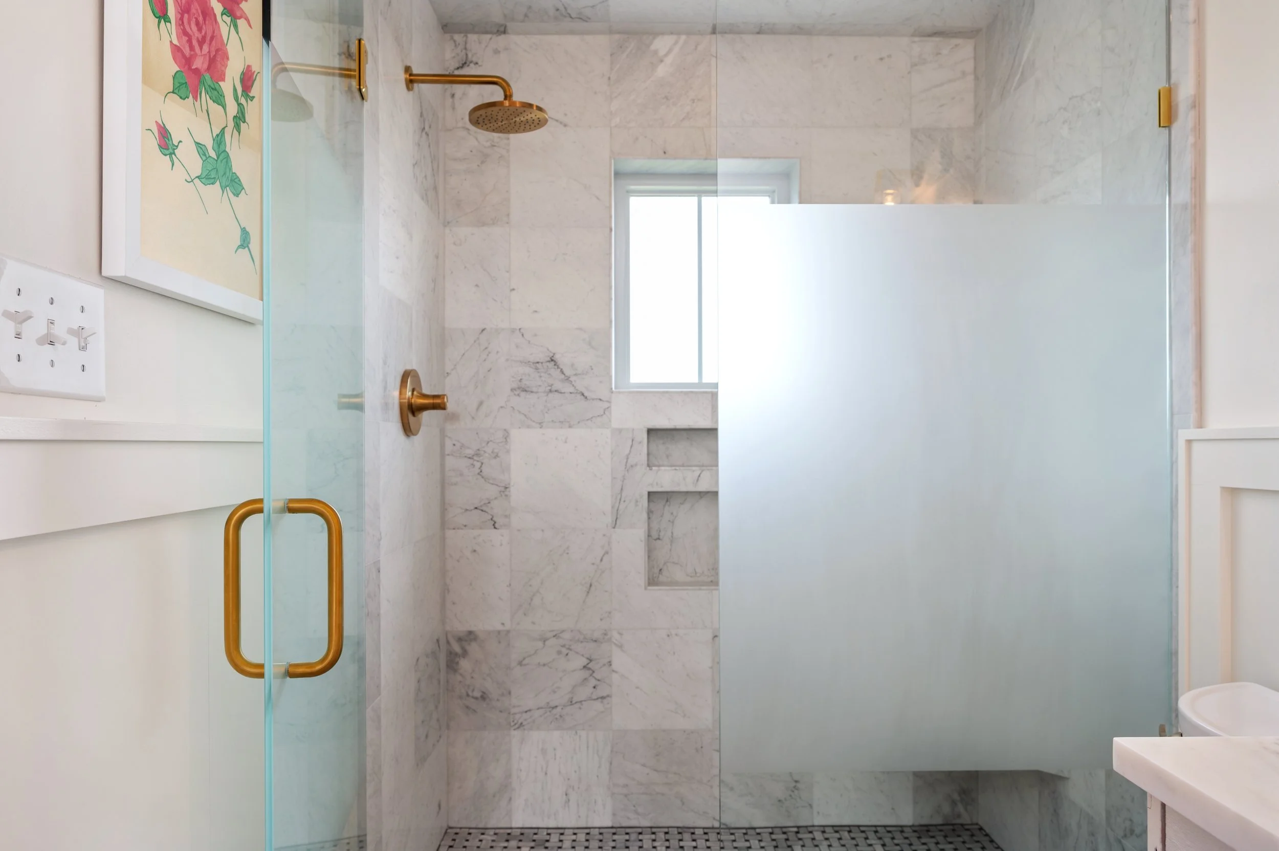Modern bathroom with marble shower walls, gold fixtures, a frosted glass door, and a small window.