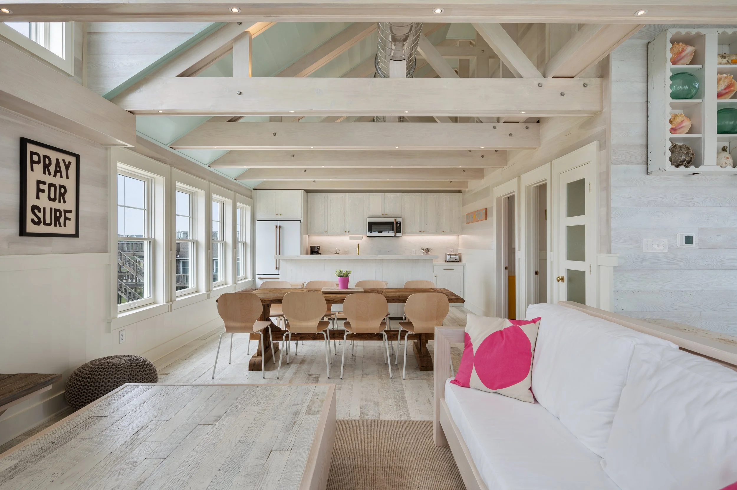 Open-concept living and kitchen area with whitewashed wood walls and exposed ceiling beams, multiple windows, a wooden dining table with eight chairs, a white sofa with pink and white pillows, and kitchen appliances in the background.