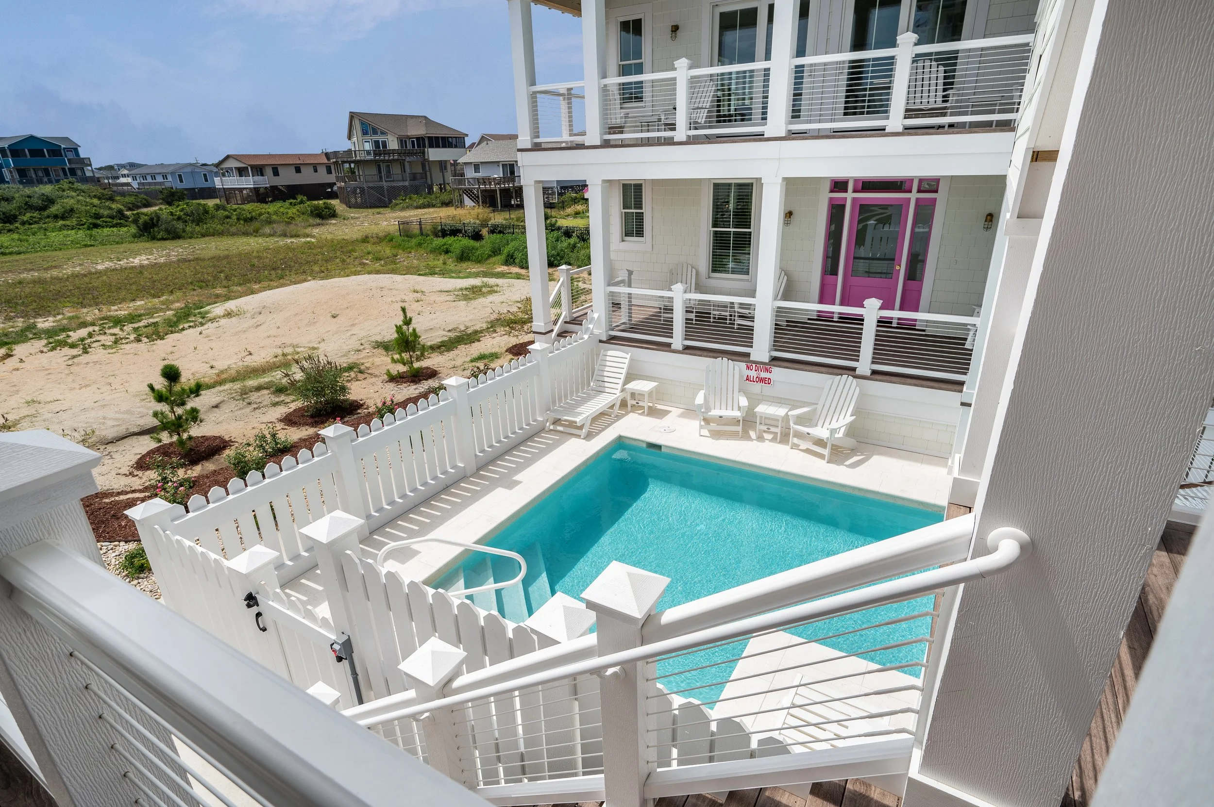 View of a small private swimming pool enclosed by a white fence, with a white house with multiple balconies in the background, and neighboring houses on a sandy, grassy landscape under a partly cloudy sky.