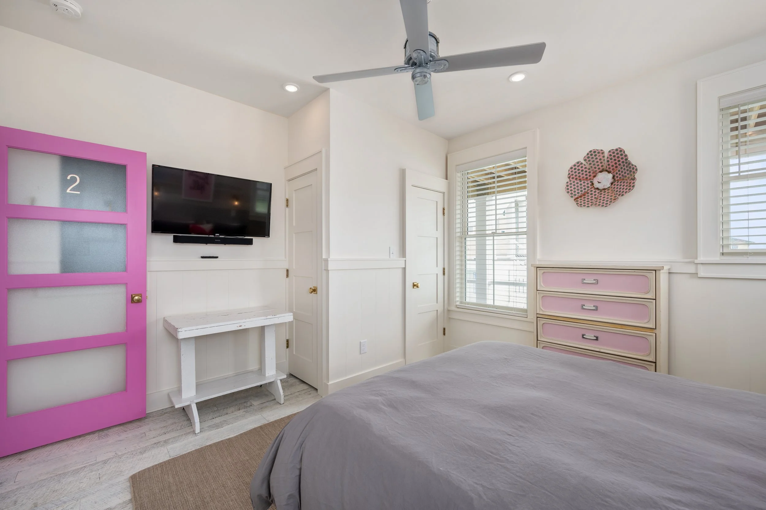 A bedroom with white walls, a ceiling fan, a flat-screen TV mounted on the wall, a pink door, a white side table, a dresser with pink drawers, and two windows with blinds, with part of a bed at the bottom of the image.