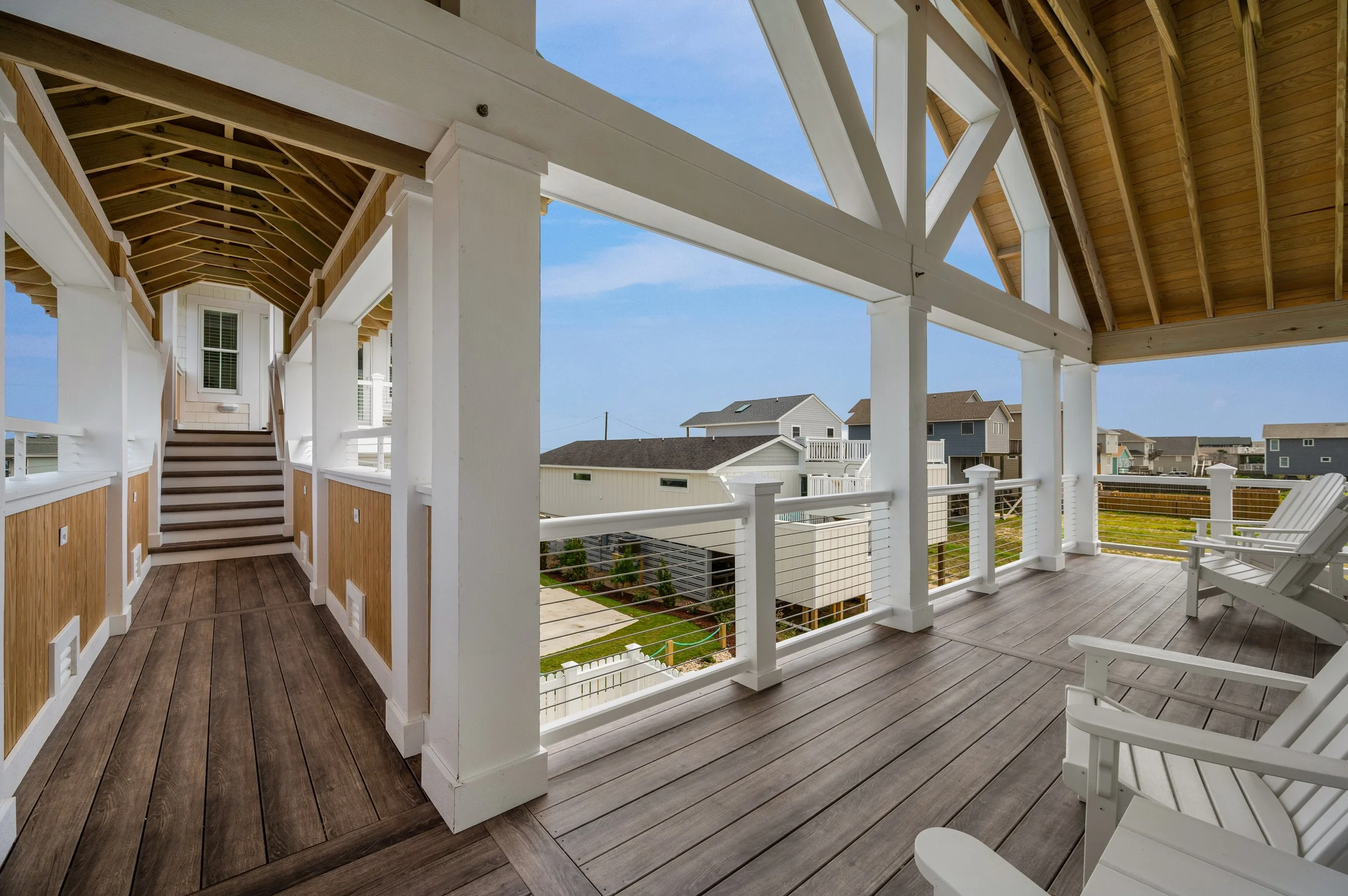 View of a spacious wooden porch with white railings and Adirondack chairs, overlooking neighboring houses under a blue sky.