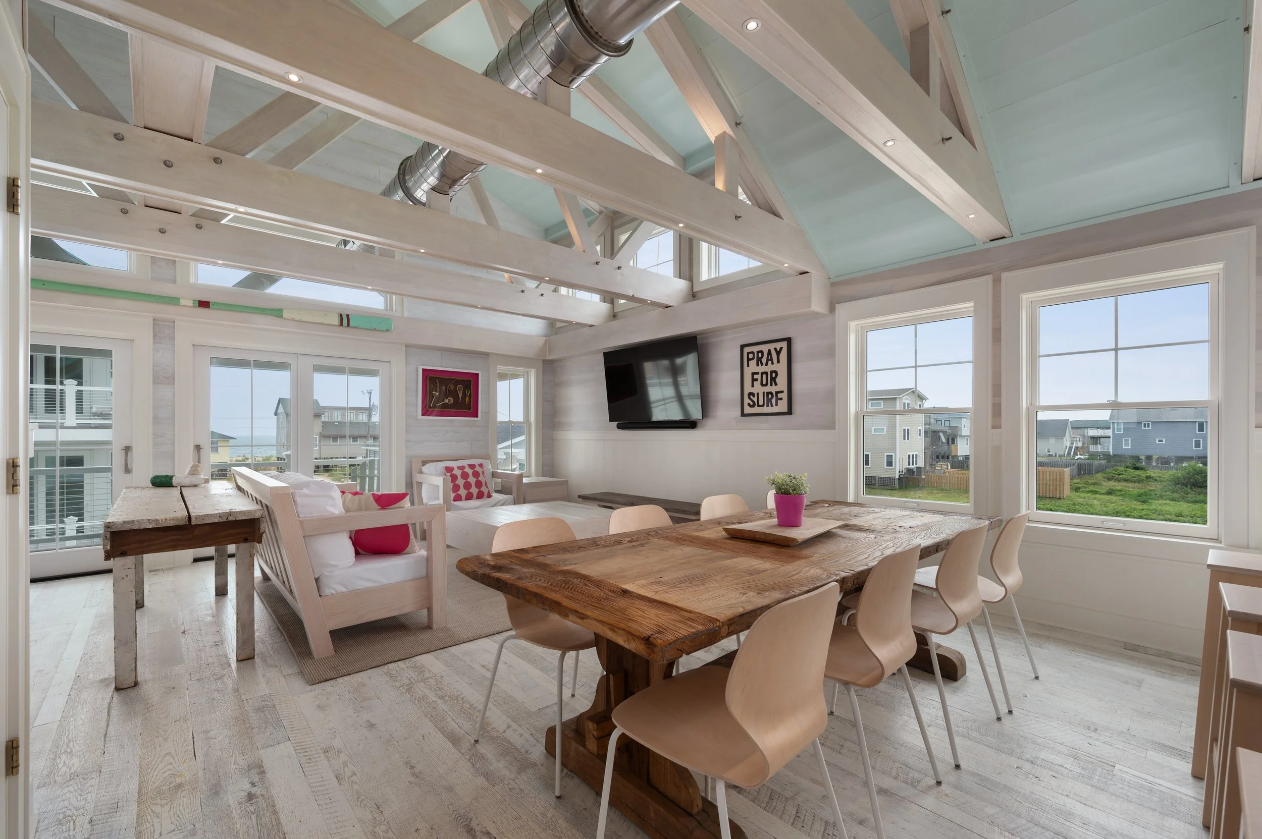 Open living and dining area in a beach house with large windows, wooden beams, and light-colored furniture, including a dining table with chairs, a couch, and wall decorations.