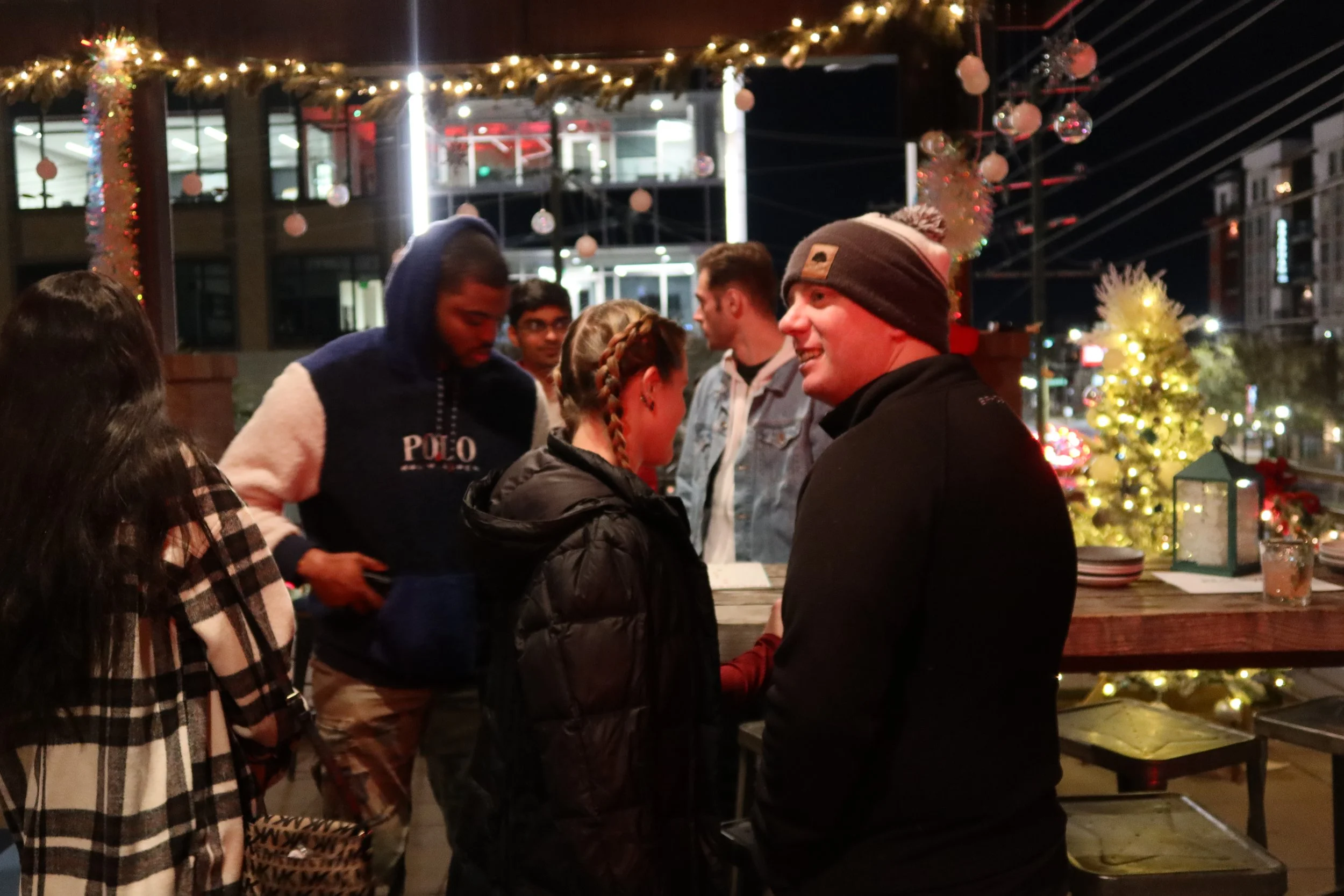 Group of people enjoying a festive outdoor gathering at night, decorated with Christmas ornaments, lights, and a Christmas tree.