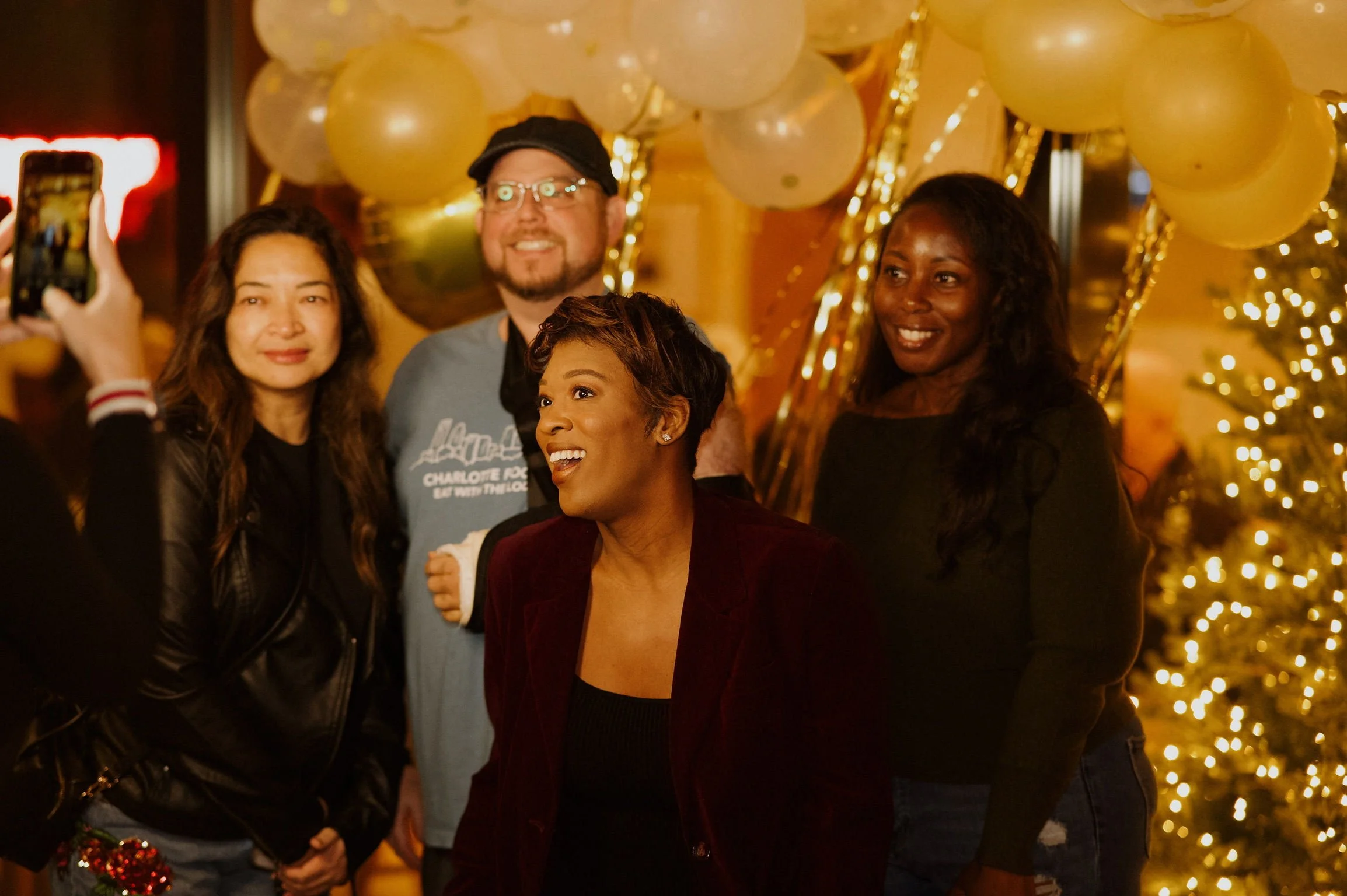 Four people celebrating at a festive party with gold balloons and Christmas lights in the background. One woman with short hair and a maroon blazer is smiling, while others are gathered around her.