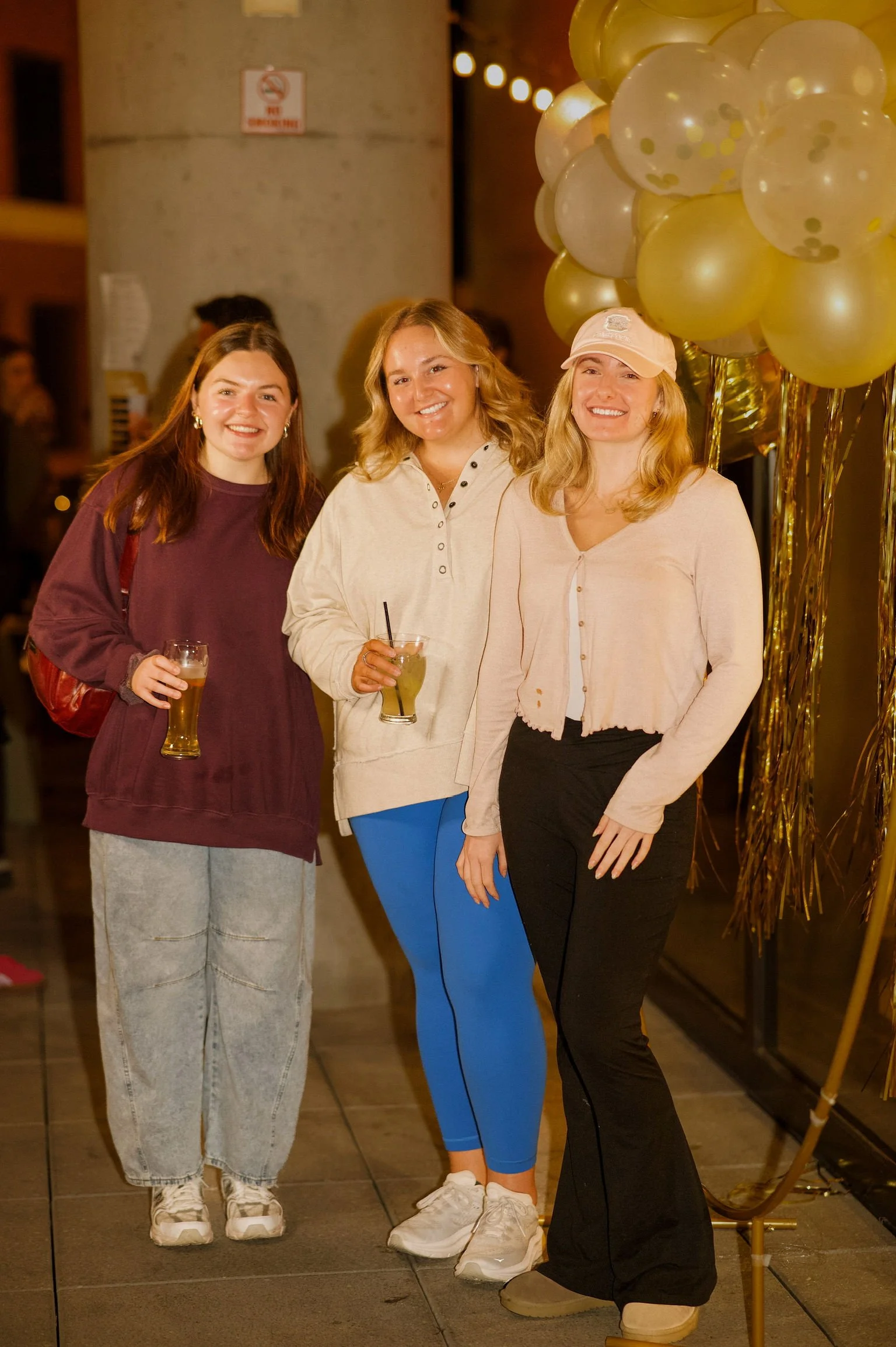 Three young women smiling and holding drinks, standing next to a bunch of gold and clear balloons, at a party or celebration.