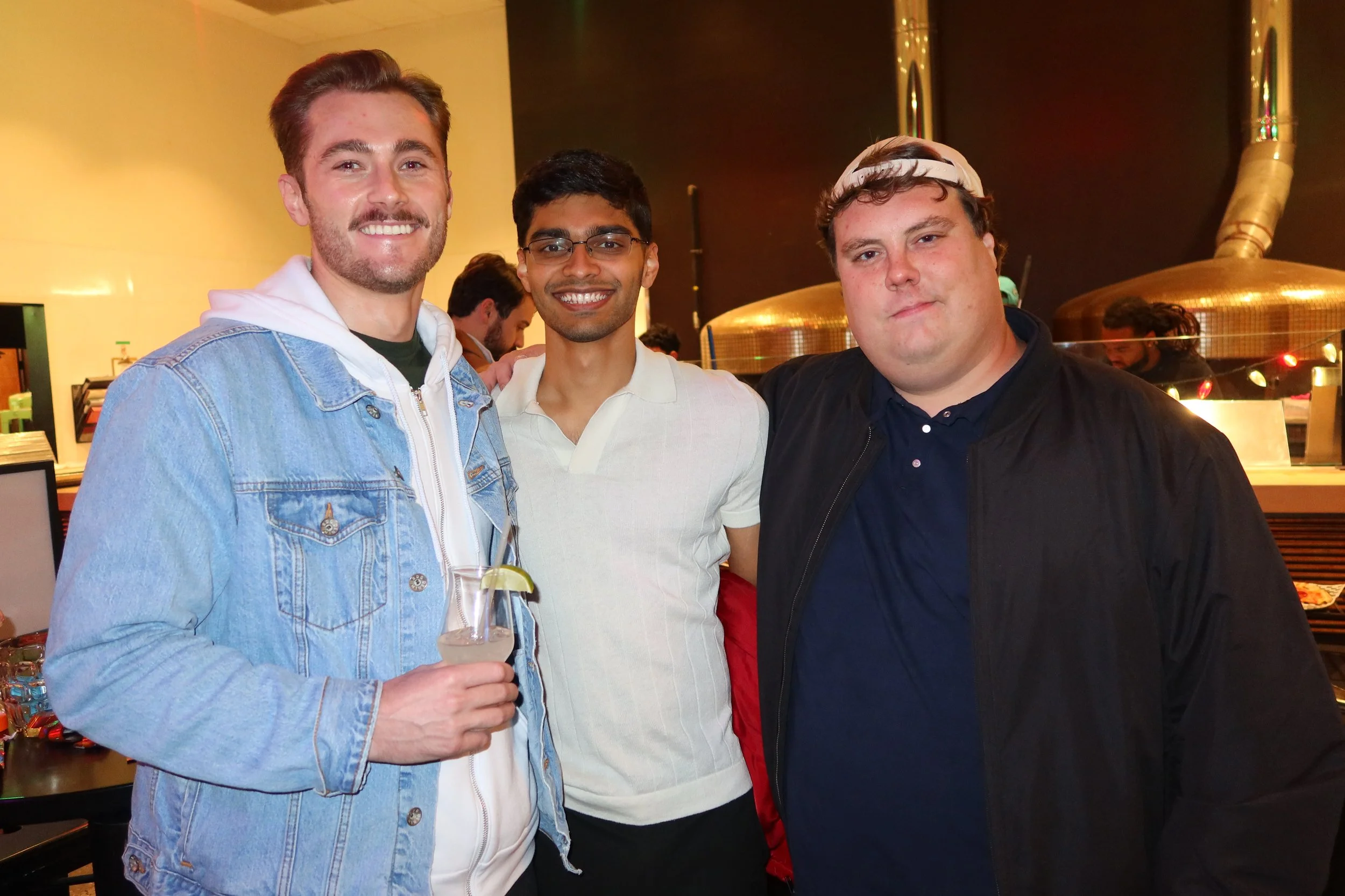 Three young men standing close together in a restaurant or bar, smiling at the camera. The man on the left holds a drink with a lemon wedge, wearing a denim jacket over a hoodie. The man in the middle wears glasses and a white polo shirt. The man on 