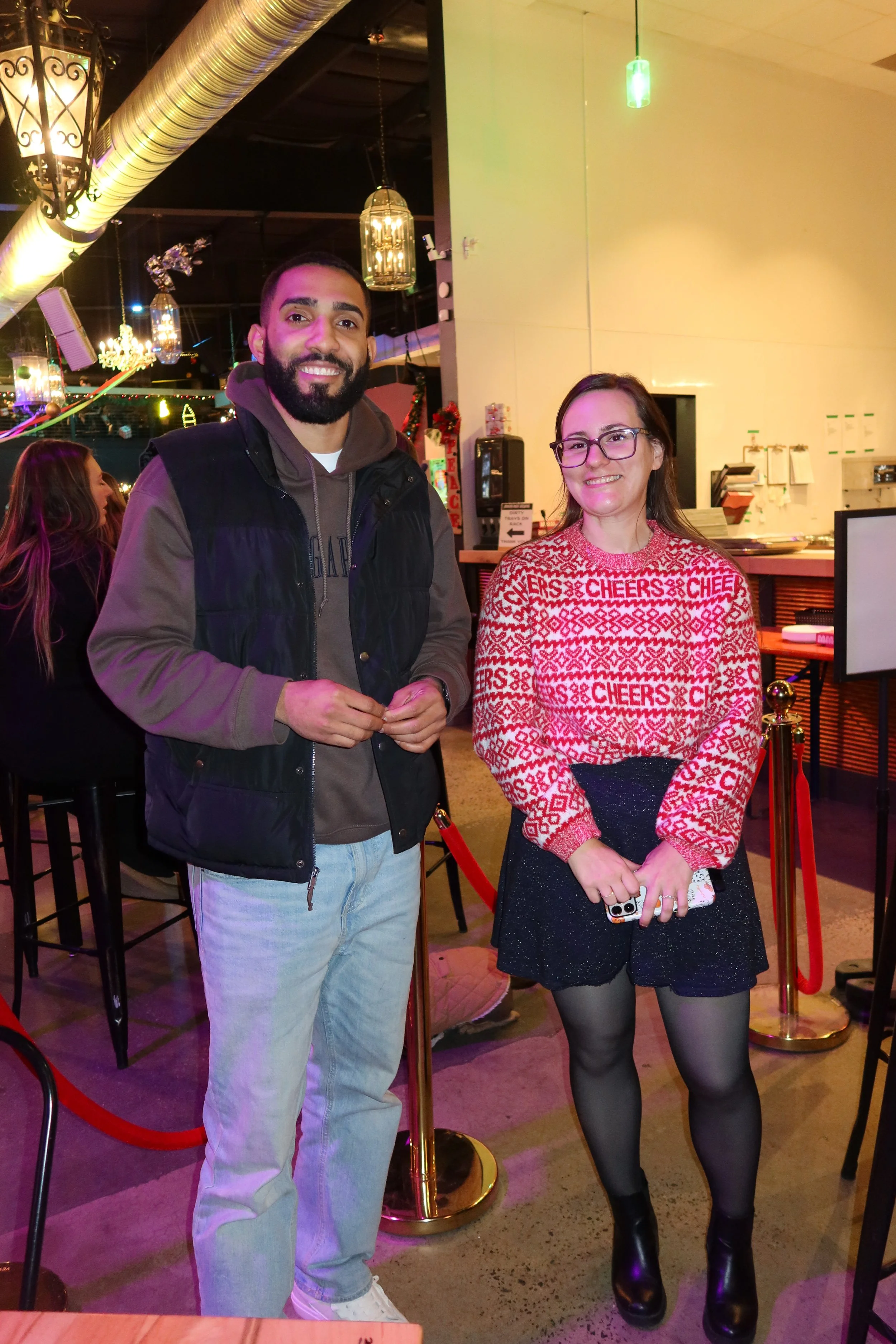 A man and a woman standing together in a festive indoor setting, smiling at the camera. The woman is wearing a red and white Christmas sweater with the word 'CHEERS' repeatedly printed on it, a black skirt, tights, and black boots. The man is wearing