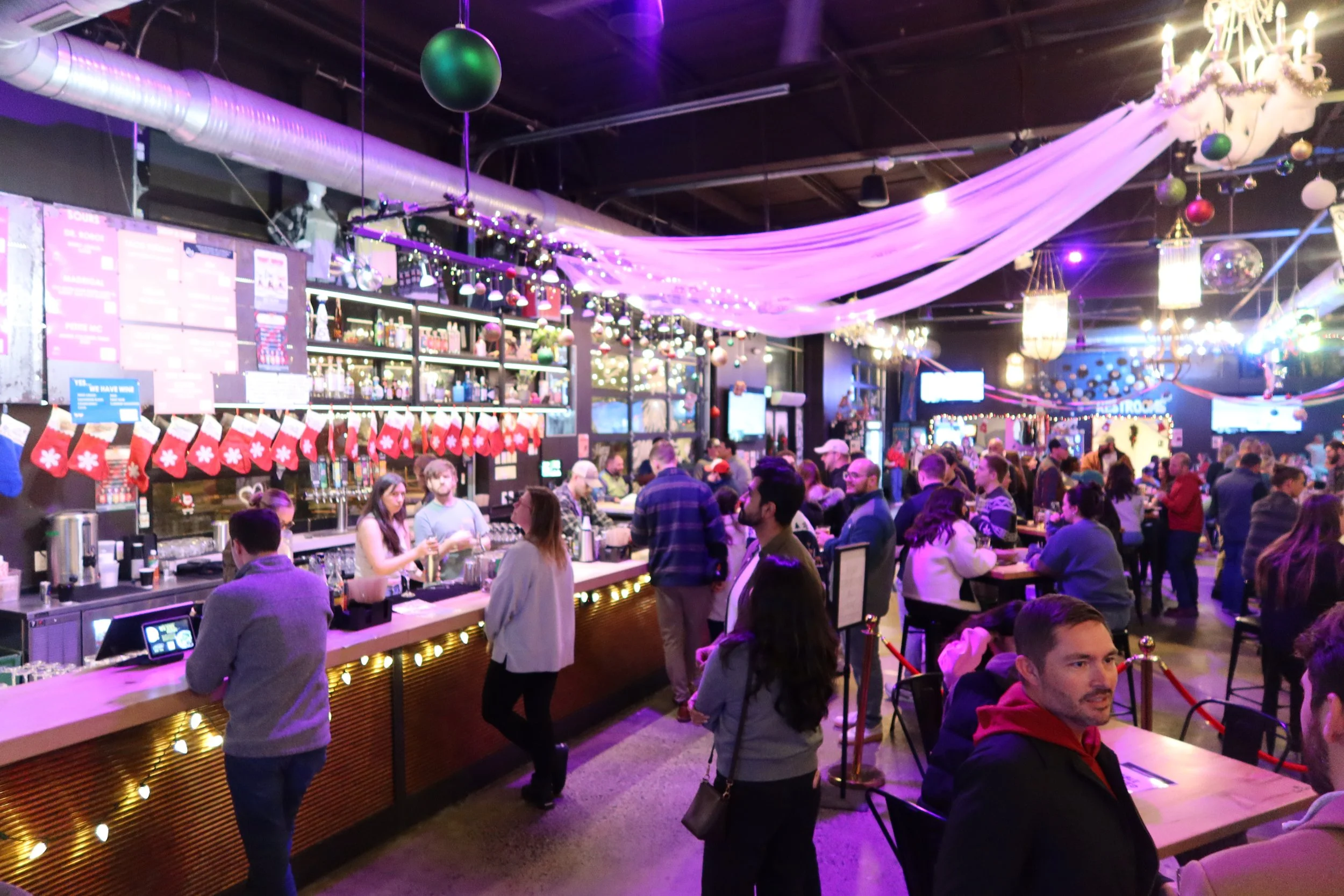 A crowded bar with people socializing and standing around the counter. The bar is decorated with Christmas stockings and holiday ornaments. The ceiling has draped pink fabric and hanging Christmas ornaments. There are several screens and illuminated 