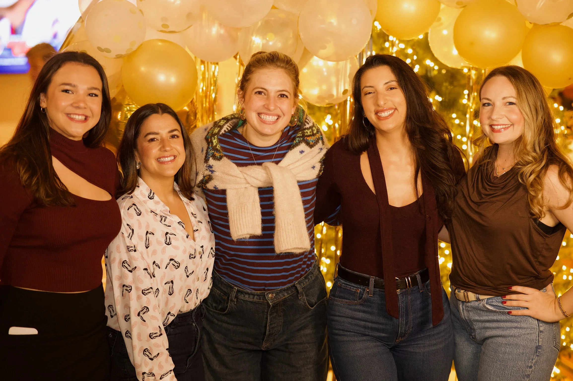 Group of five women smiling at a celebration with gold balloons and string lights in the background.