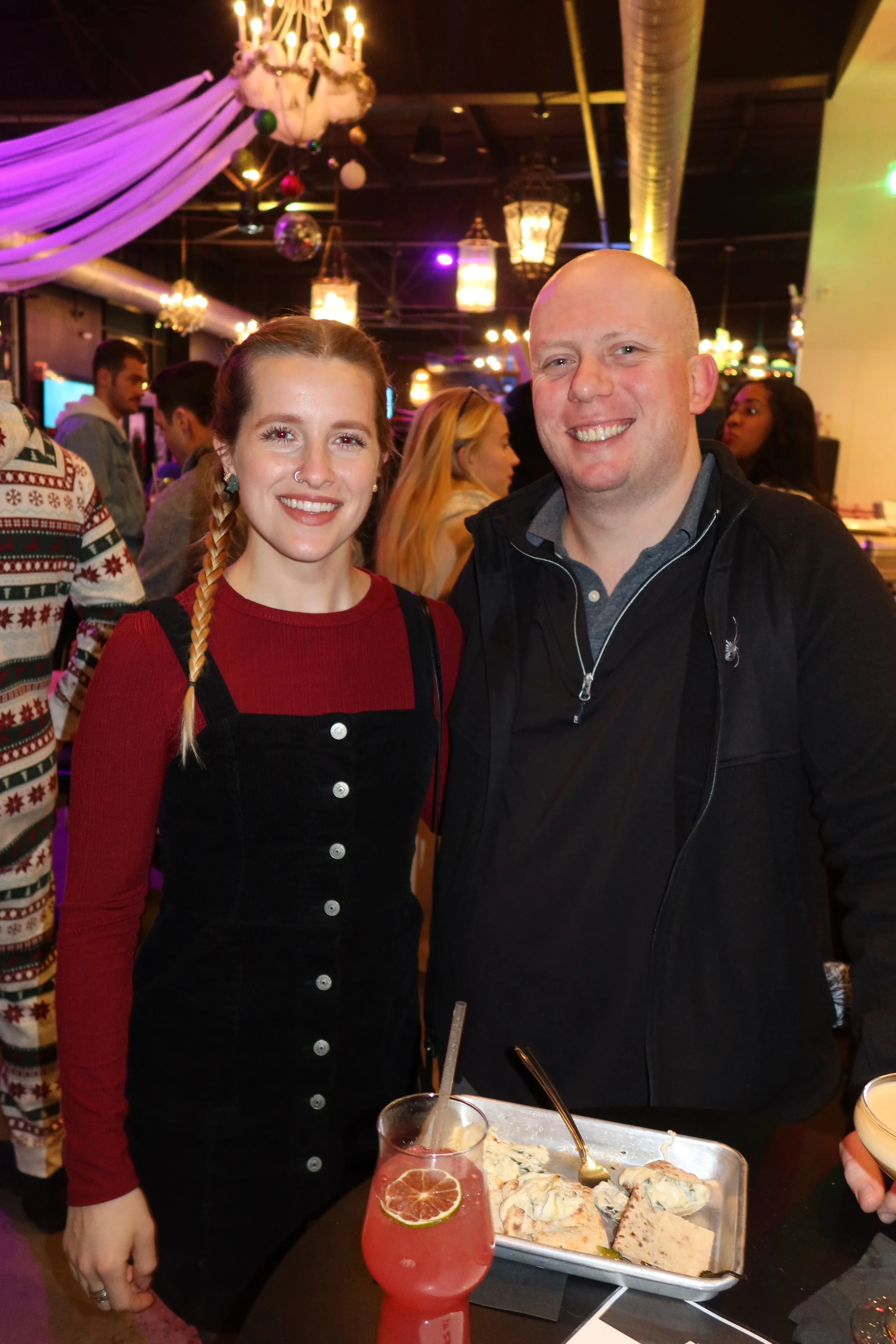 A young woman with a braid and a young man smiling at a party or social event indoors with festive lighting and decorations, a young woman in the background, and a table with a glass of pink drink and a tray of food