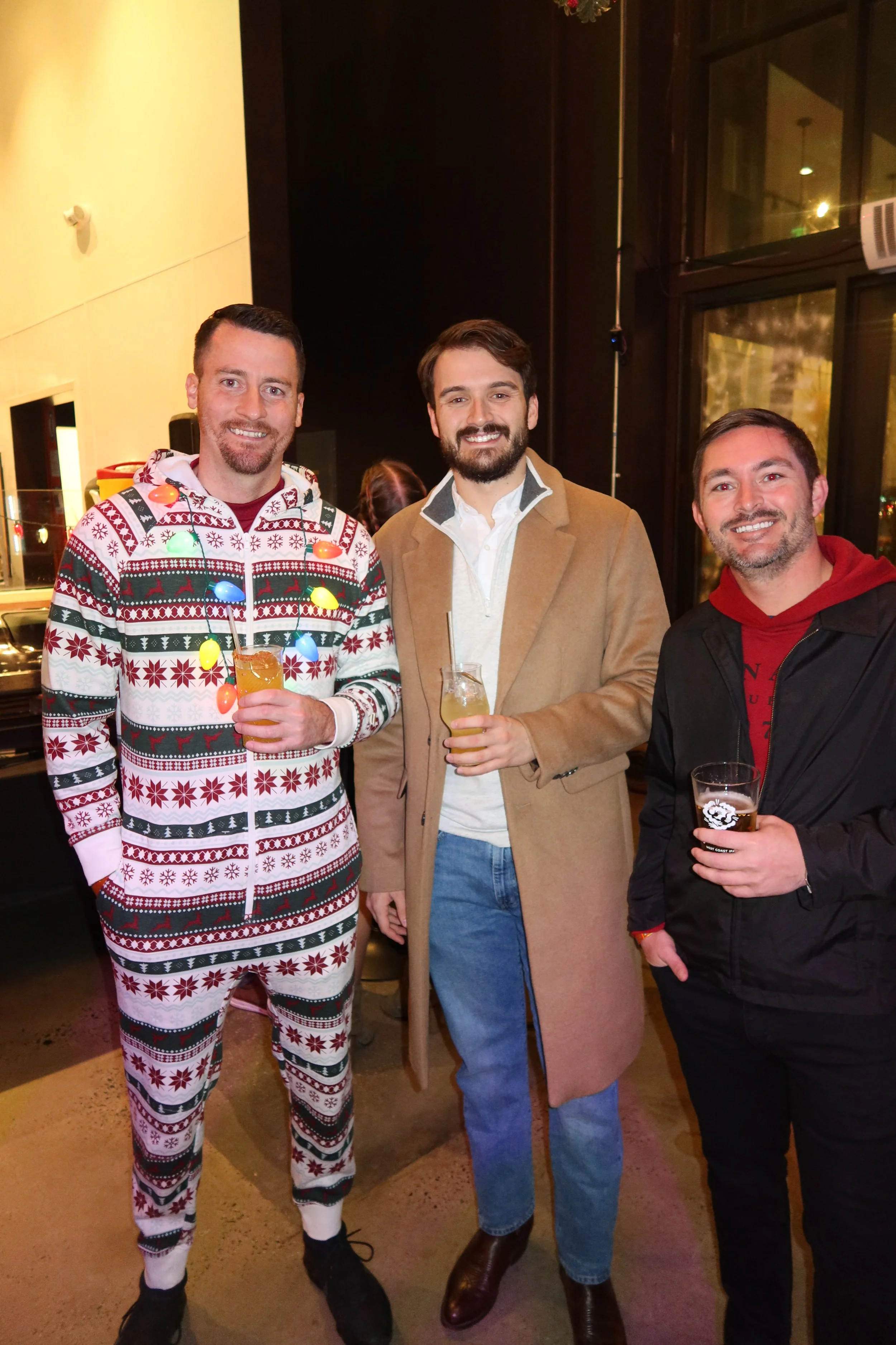 Three men standing together in a bar, smiling, holding drinks, celebrating, one wearing a holiday-themed onesie, another in a tan coat, and the third in a black jacket.