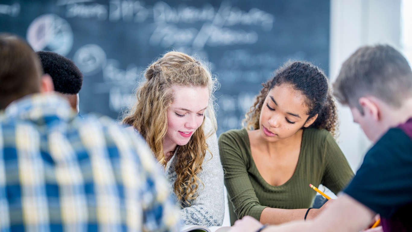 Group of students studying together in a classroom. Two girls in the foreground are focused on their work with one holding a pencil, while students in the background and to the sides are engaged in their tasks.