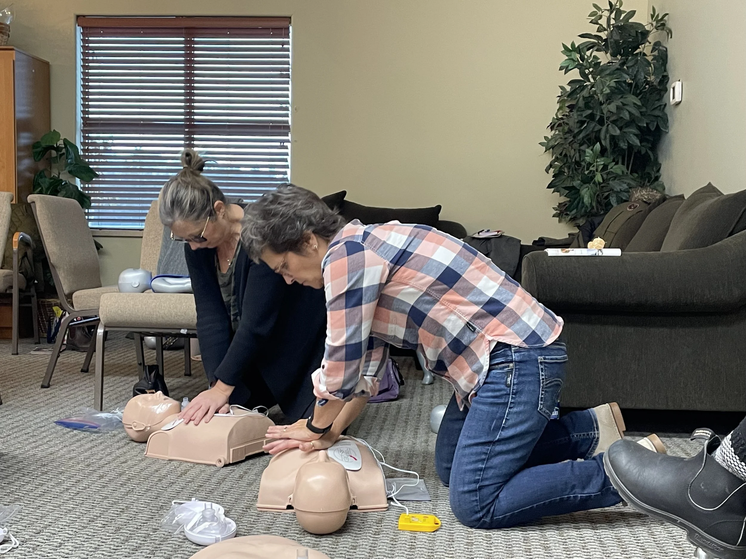 Two women practicing CPR on mannequins during a training session in a room with chairs, a sofa, and a window with blinds.