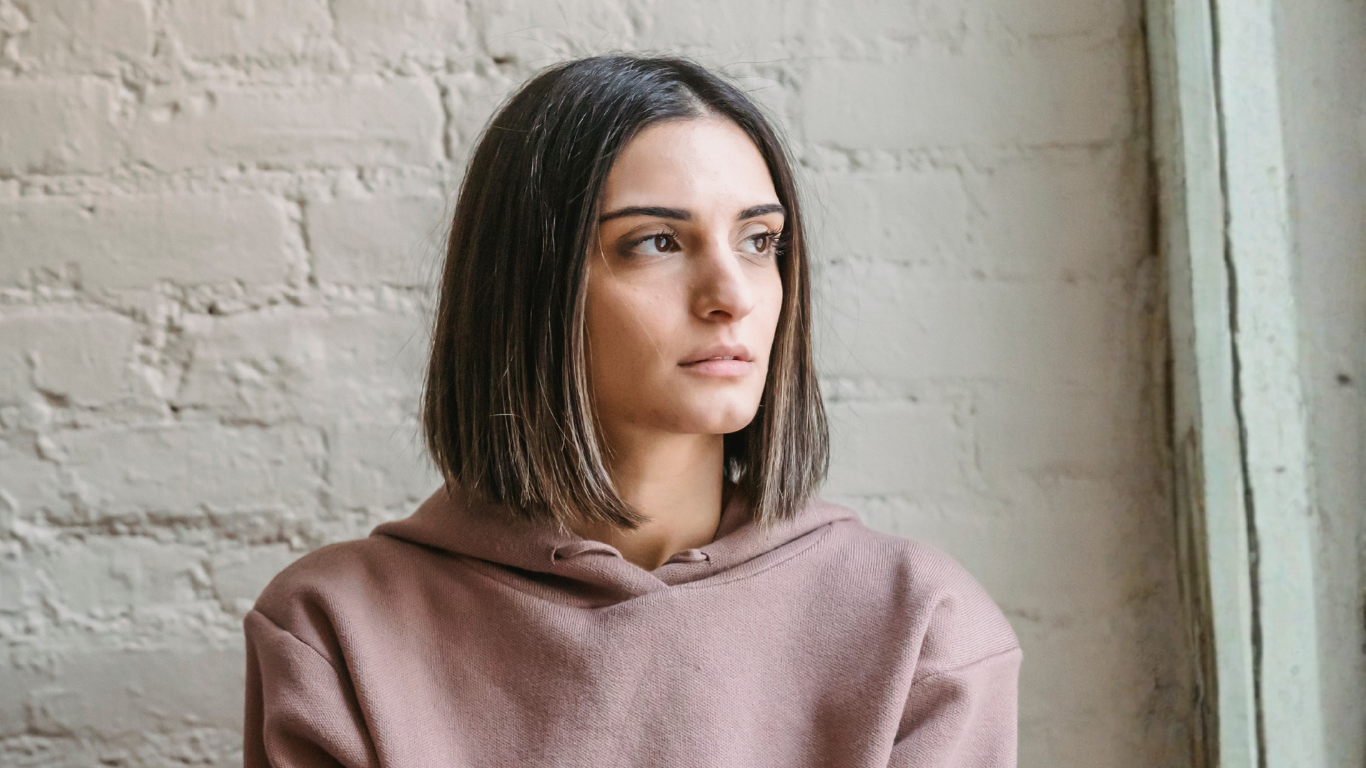 A young woman with shoulder-length brown hair is looking to her left. She is wearing a pink hoodie and is standing against a white brick wall.