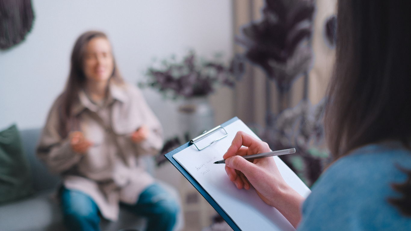 Therapist taking notes while speaking with a young woman during a therapy session in a cozy room with plants and wall decorations.