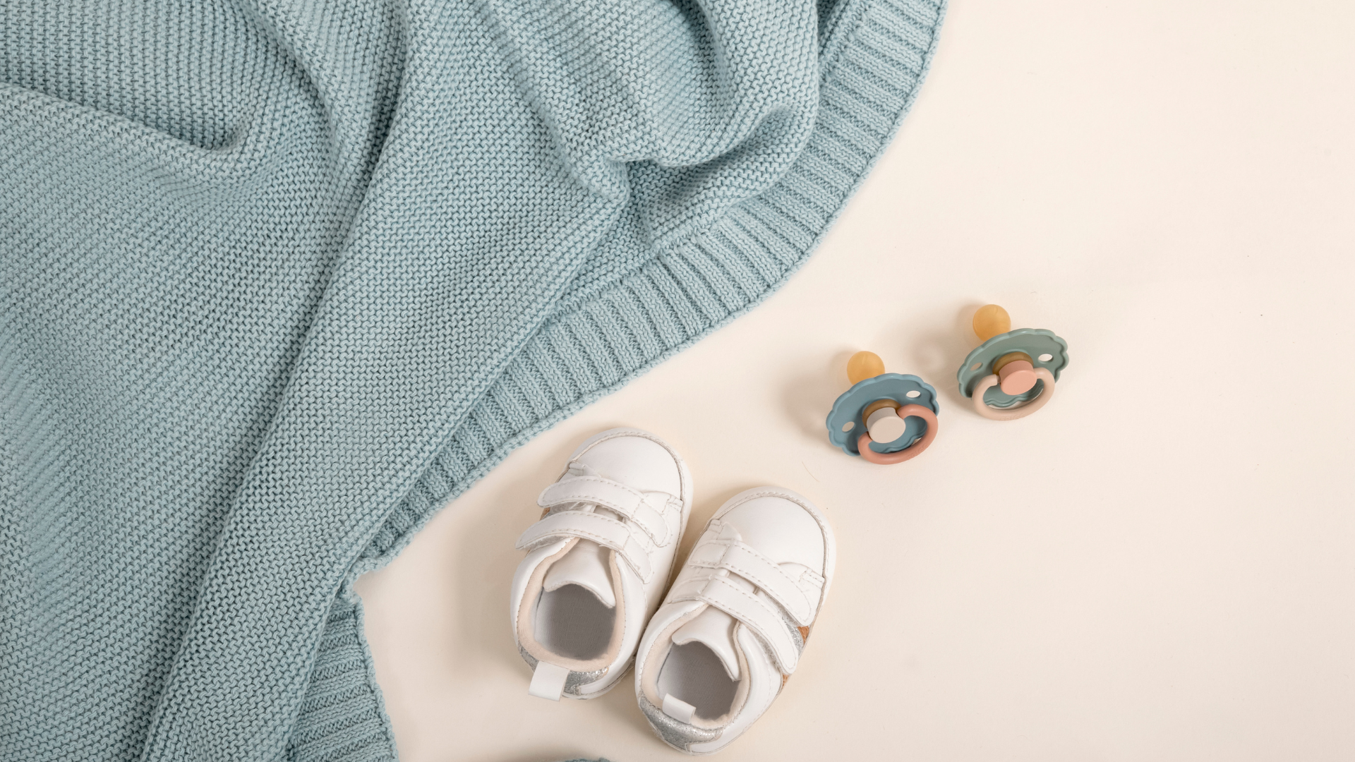 Light blue knitted blanket, white baby shoes with velcro straps, and teal and beige pacifiers on a white surface.