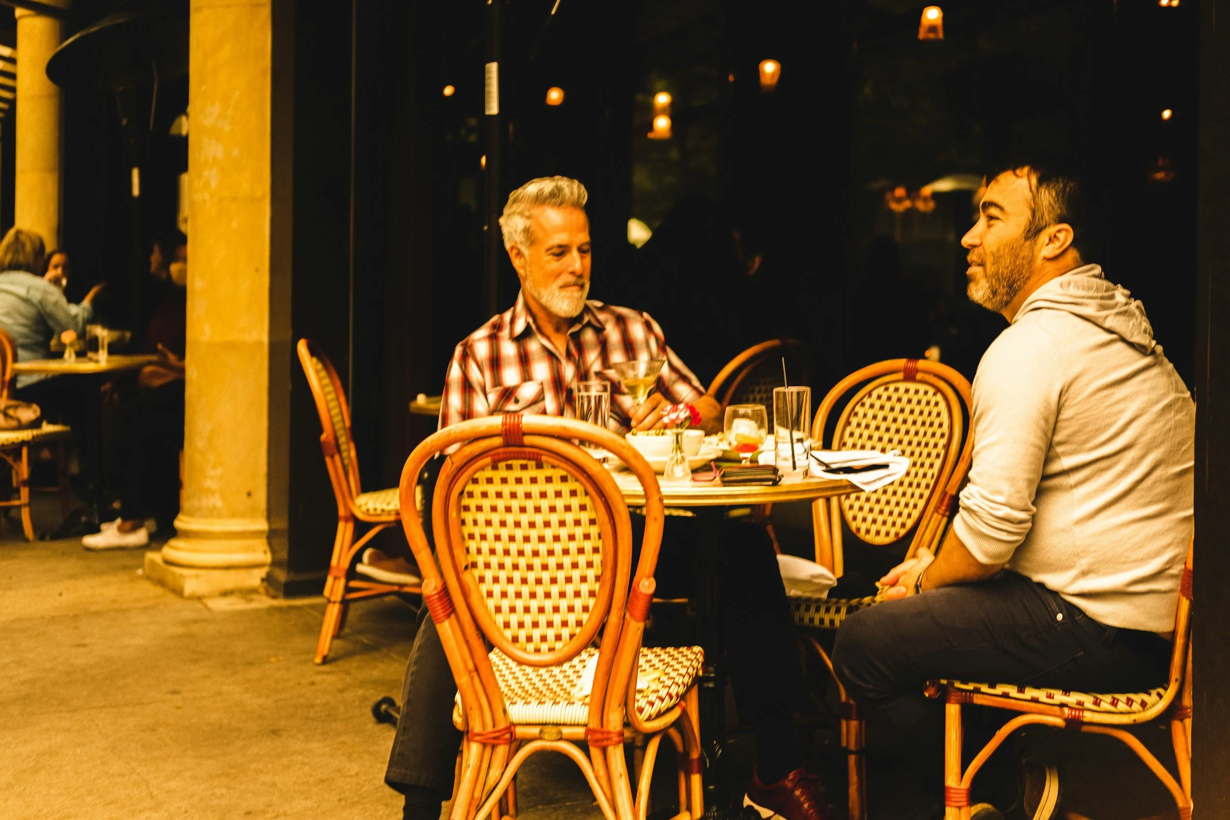Two men sitting at an outdoor restaurant table having a conversation, with drinks and small plates on the table, illuminated by warm yellow lighting at night.