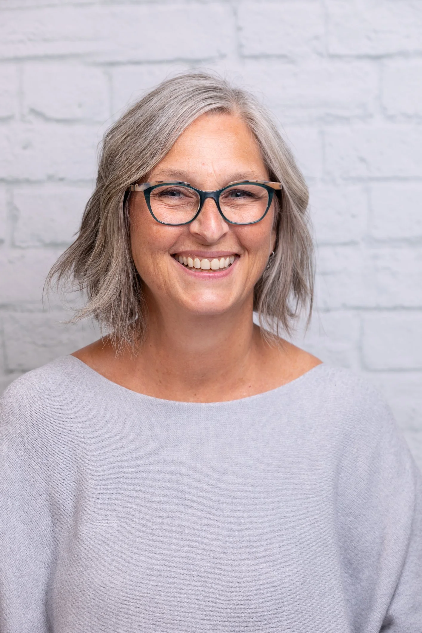 A woman with short gray hair, wearing glasses and a light gray top, smiling in front of a white brick wall.