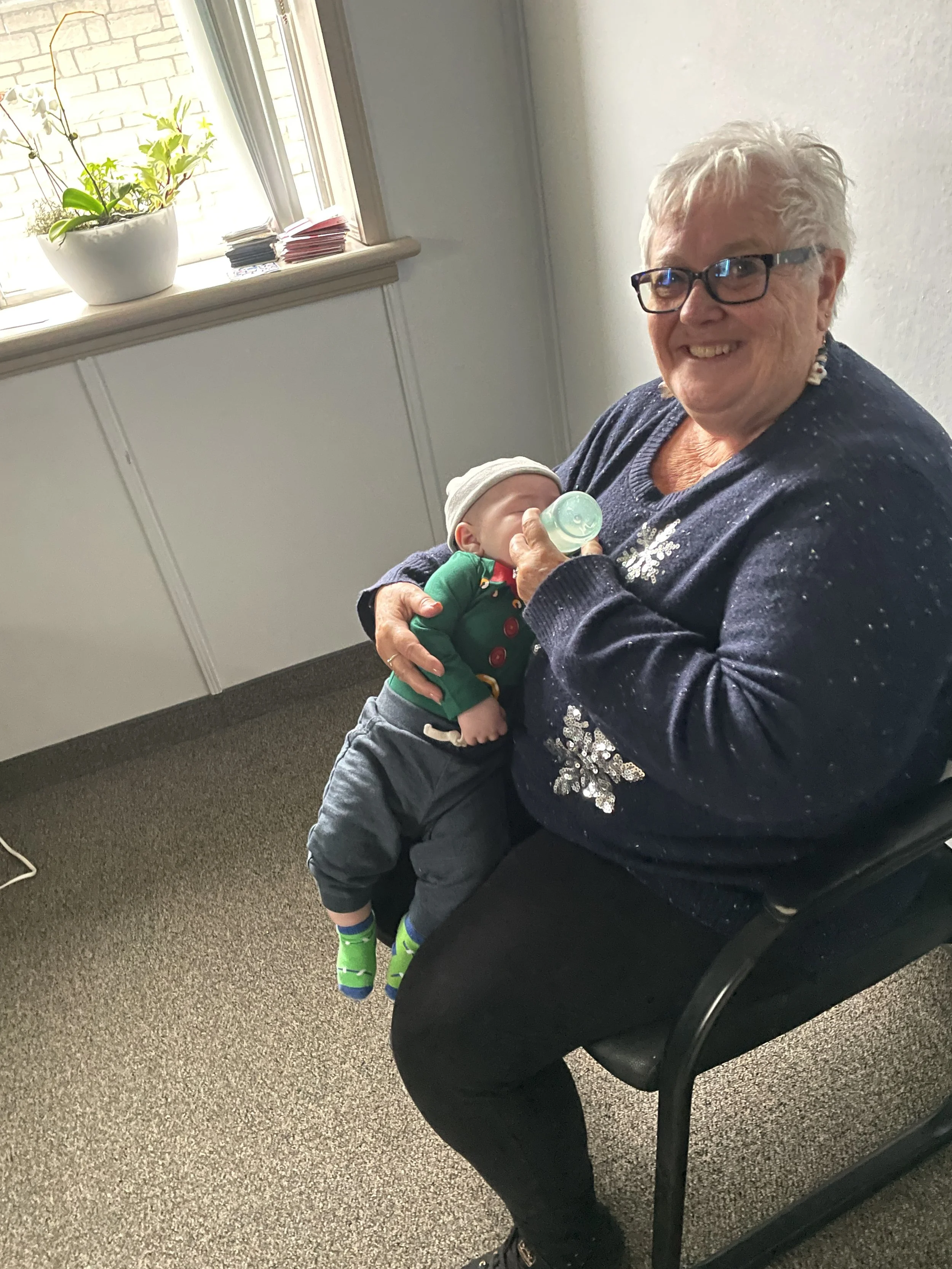 An elderly woman is sitting in a room with a baby drinking from a bottle. The woman is smiling and wearing glasses and a dark sweater with sparkles. The baby is dressed in a green outfit with a beanie and colorful socks.