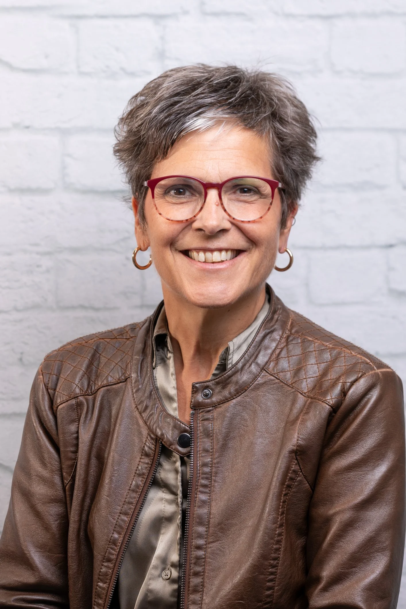 A smiling middle-aged woman with short gray hair, red glasses, hoop earrings, wearing a brown leather jacket and light-colored shirt, standing against a white brick wall.