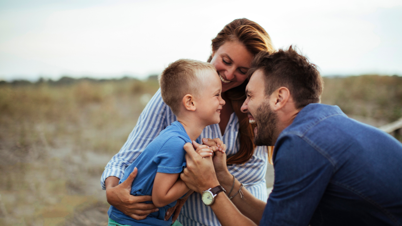 Family of three smiling and playfully fighting on a beach, with a woman, a man, and a young boy.
