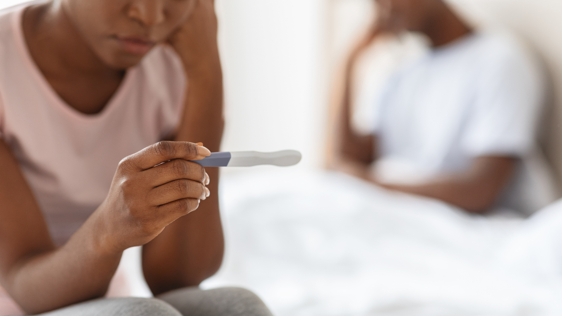 A woman holding a pregnancy test with a positive result, looking at it with concern. Another woman is in the background.