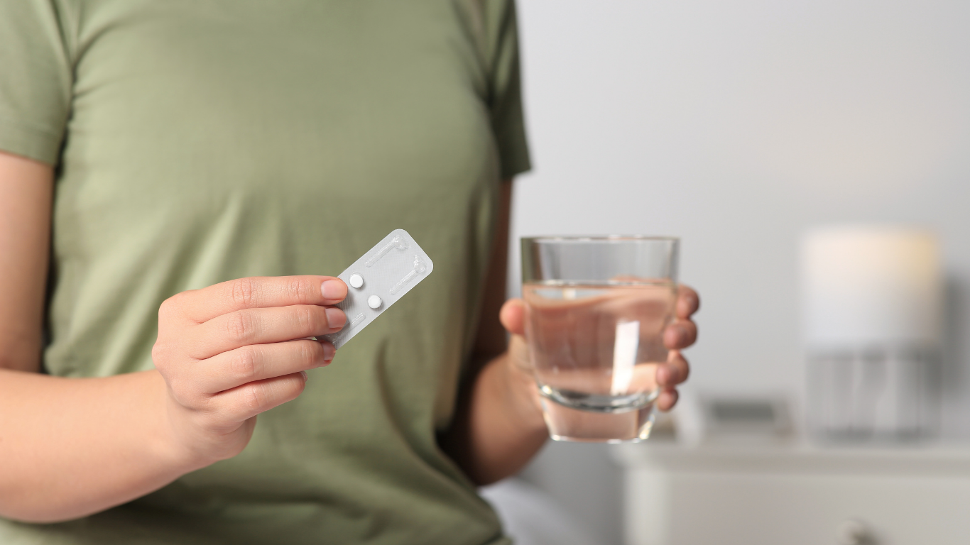 A person holding a blister pack with two pills in one hand and a glass of water in the other hand.