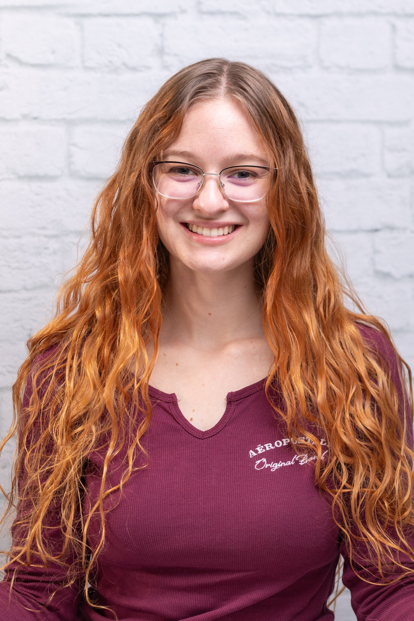 A young woman with long curly red hair, glasses, and a maroon shirt standing in front of a white brick wall.