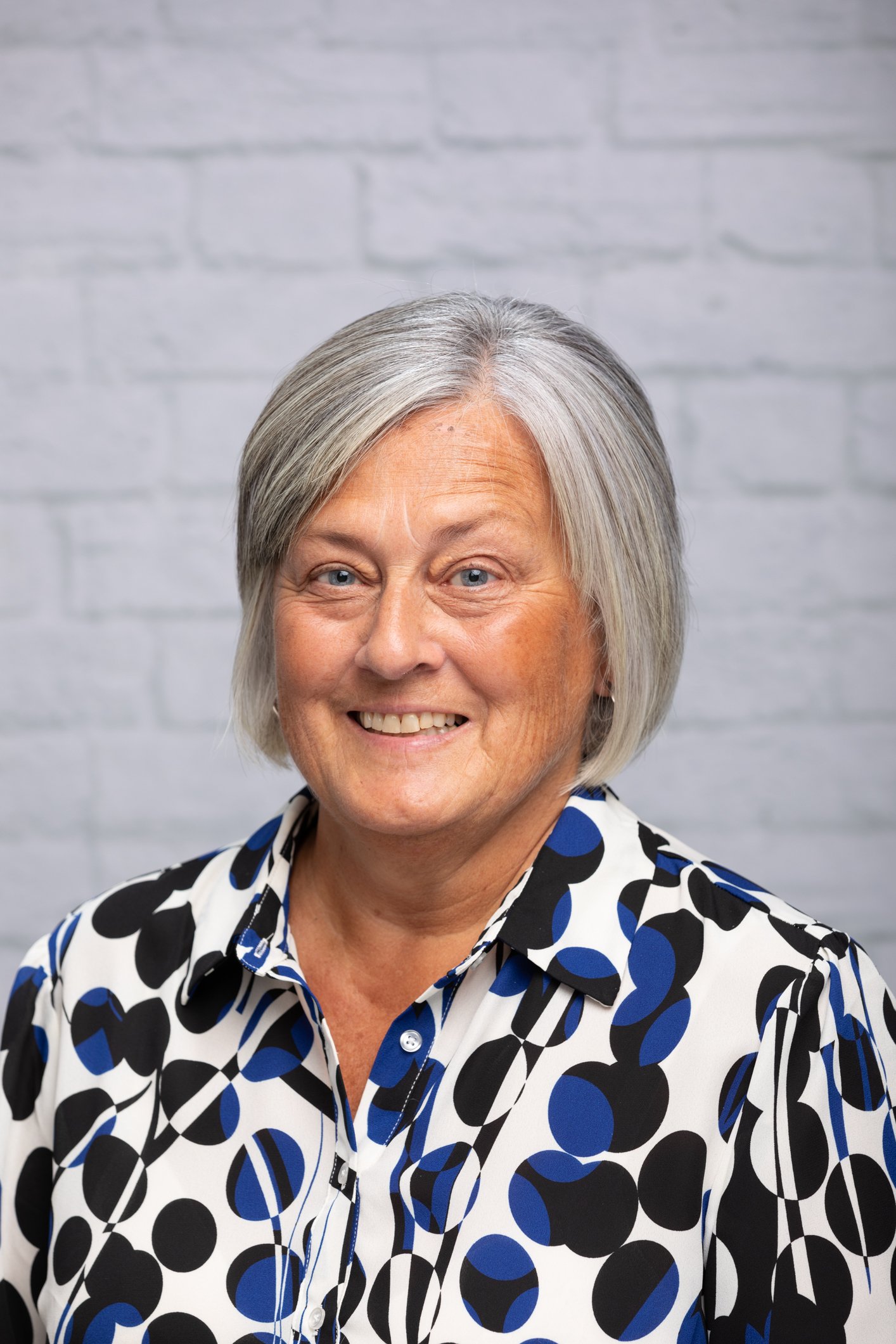 Portrait of an elderly woman with short gray hair, smiling, wearing a black, white, and blue patterned blouse, standing against a white brick wall background.