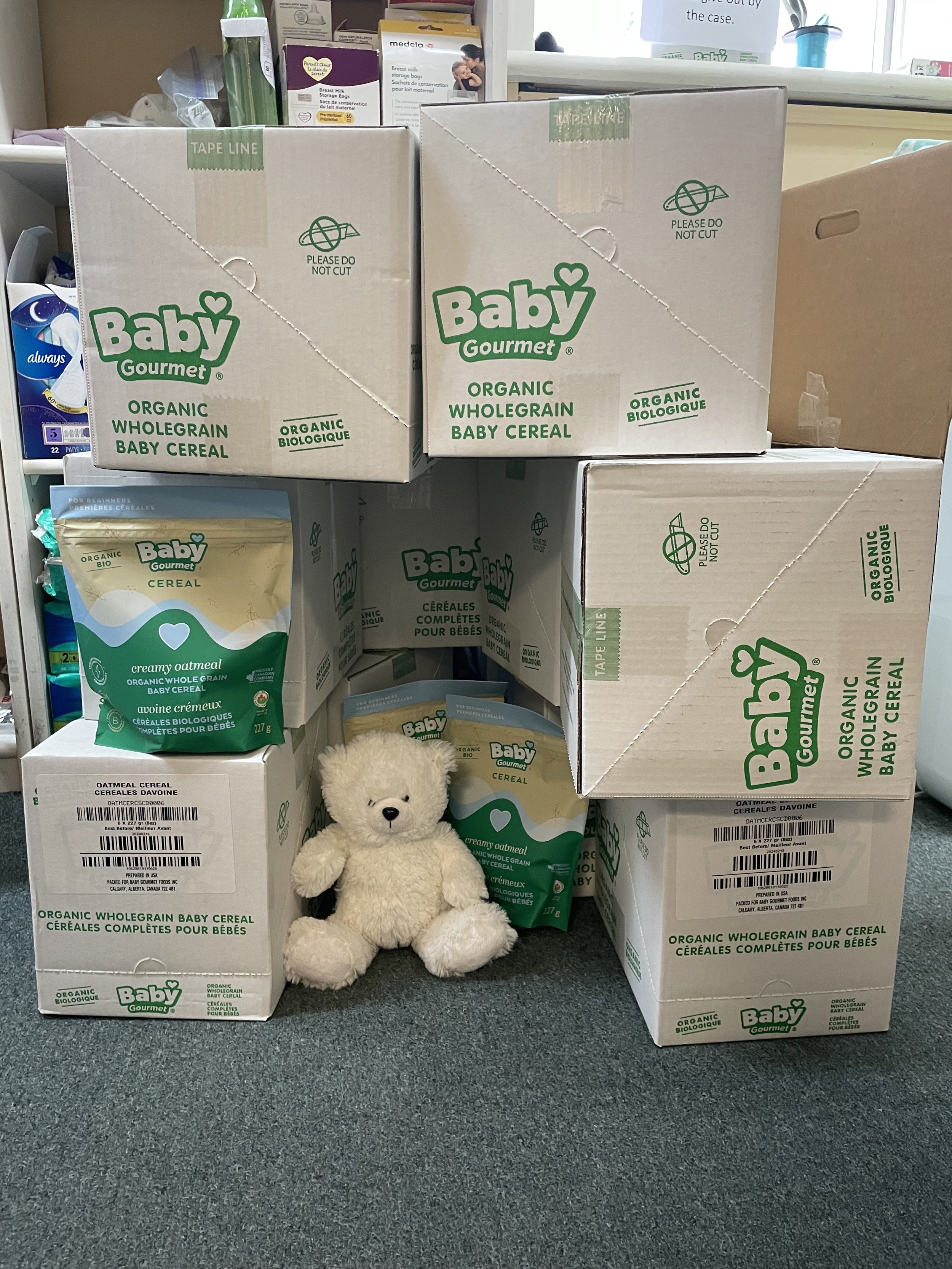 Stacks of boxes and packages of Baby Gourmet organic whole grain baby cereal, with a plush teddy bear in front on a gray carpeted floor in a store or storage area.