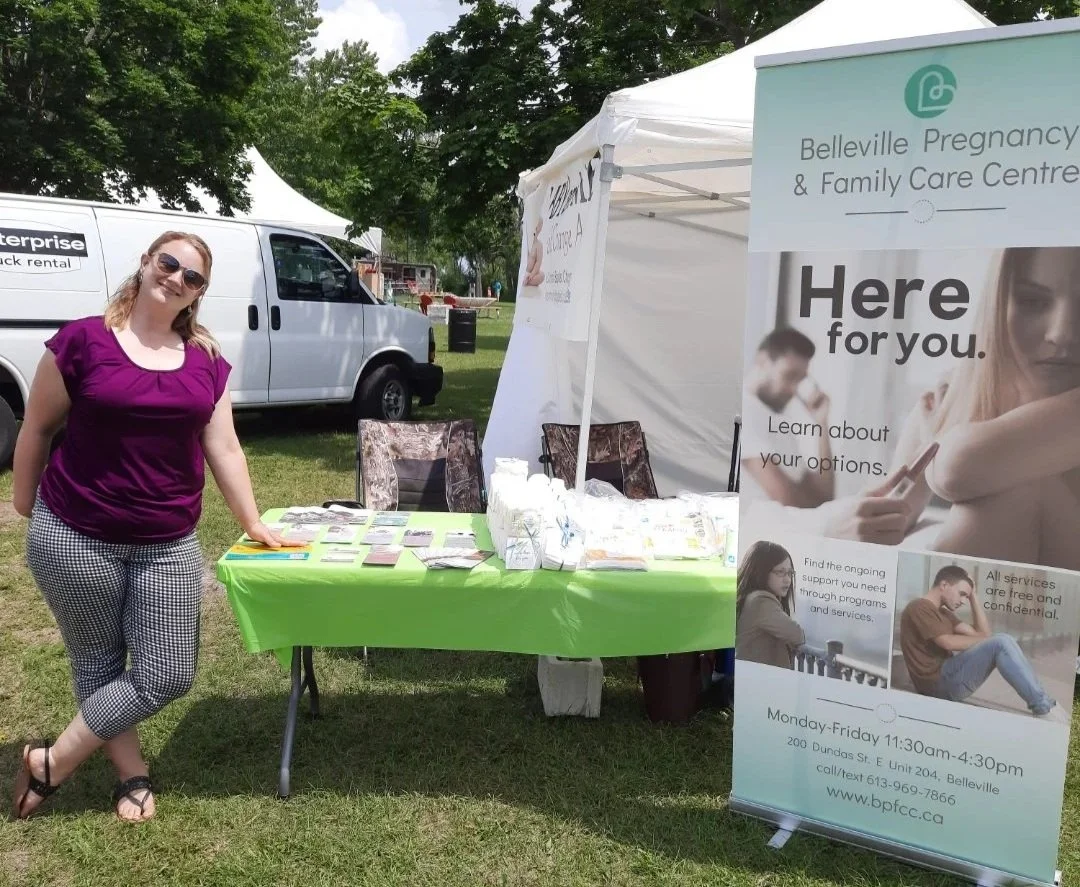Woman standing next to a table offering free diapers and supplies at an outdoor community event, with a sign for Belleville Pregnancy & Family Care Centre nearby.