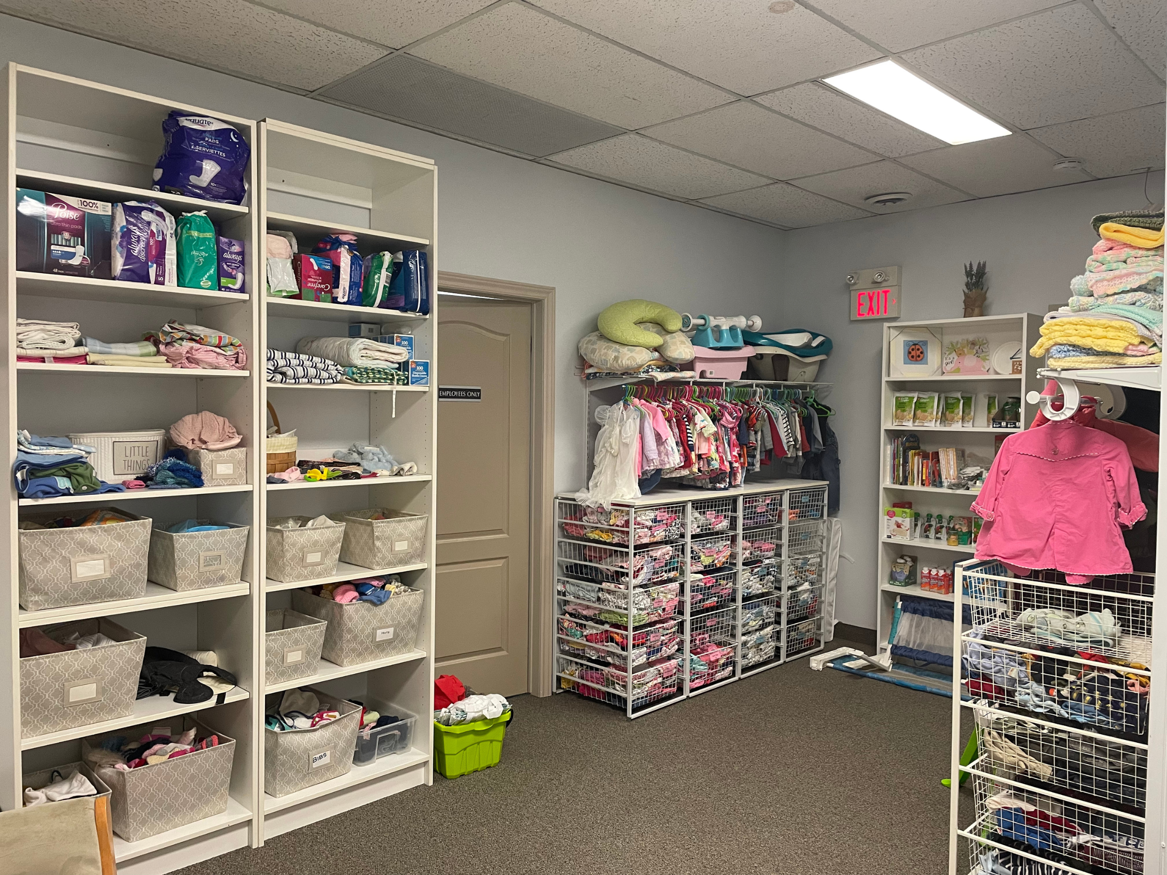 A room with shelves filled with baby clothes, diapers, and related items. There is a clothing rack with small children's outfits, a white laundry basket with clothes, and neatly folded towels on a shelf. An exit sign is visible on the back wall.
