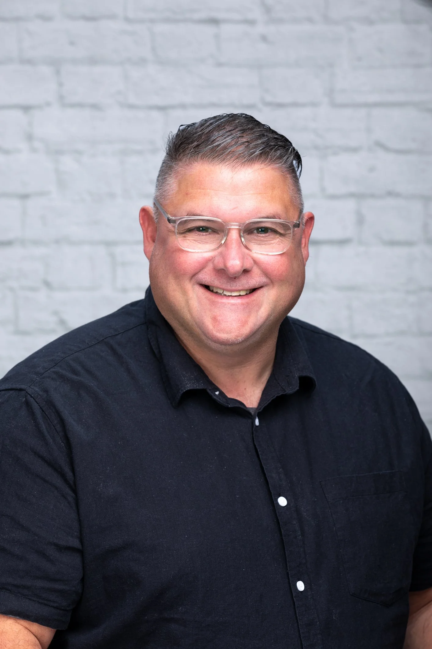 A smiling man with short gray hair, glasses, and a dark button-up shirt against a white brick wall background.