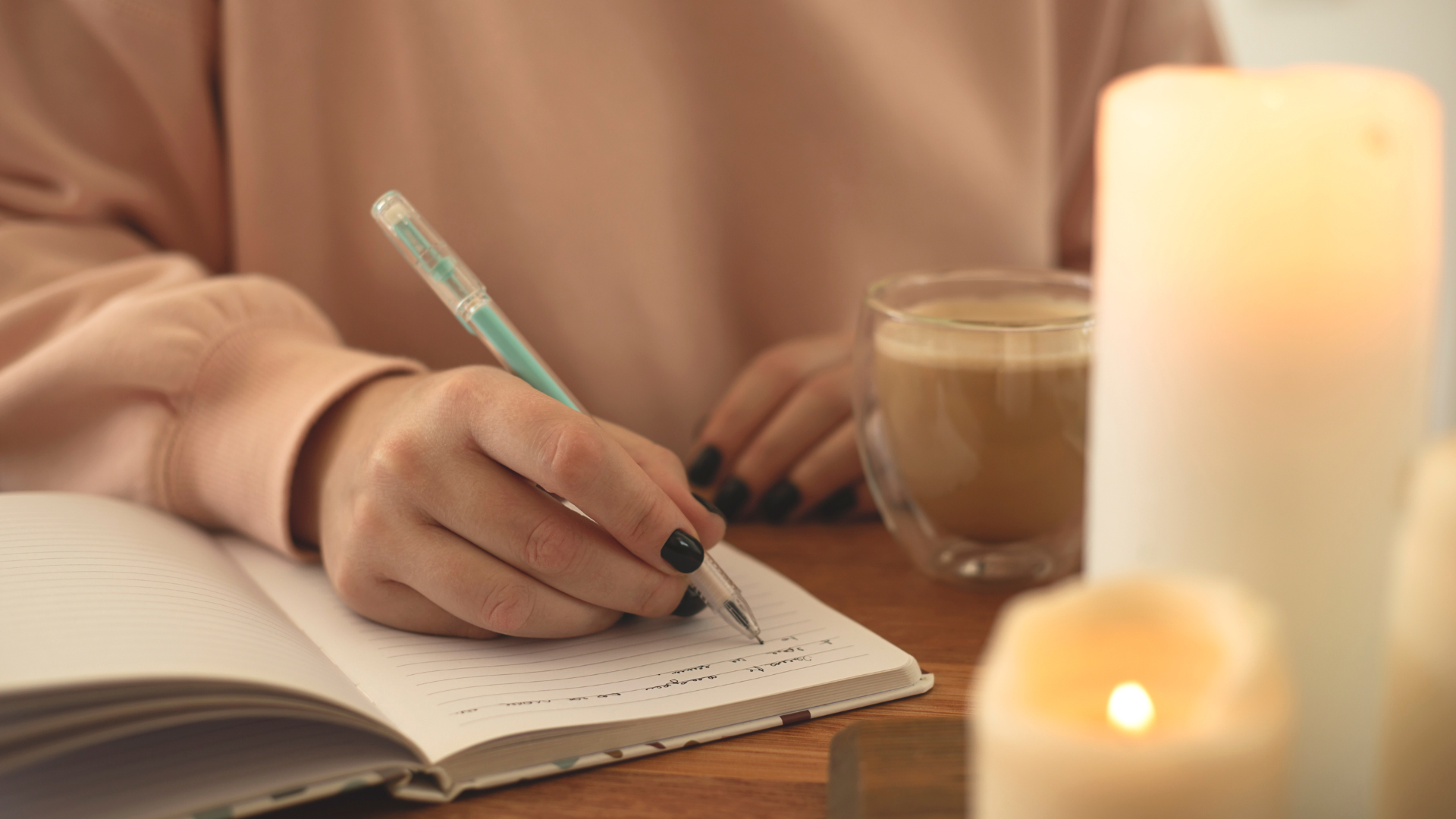 A person writing in a notebook with a pen, sitting at a wooden table with candles and a hot beverage