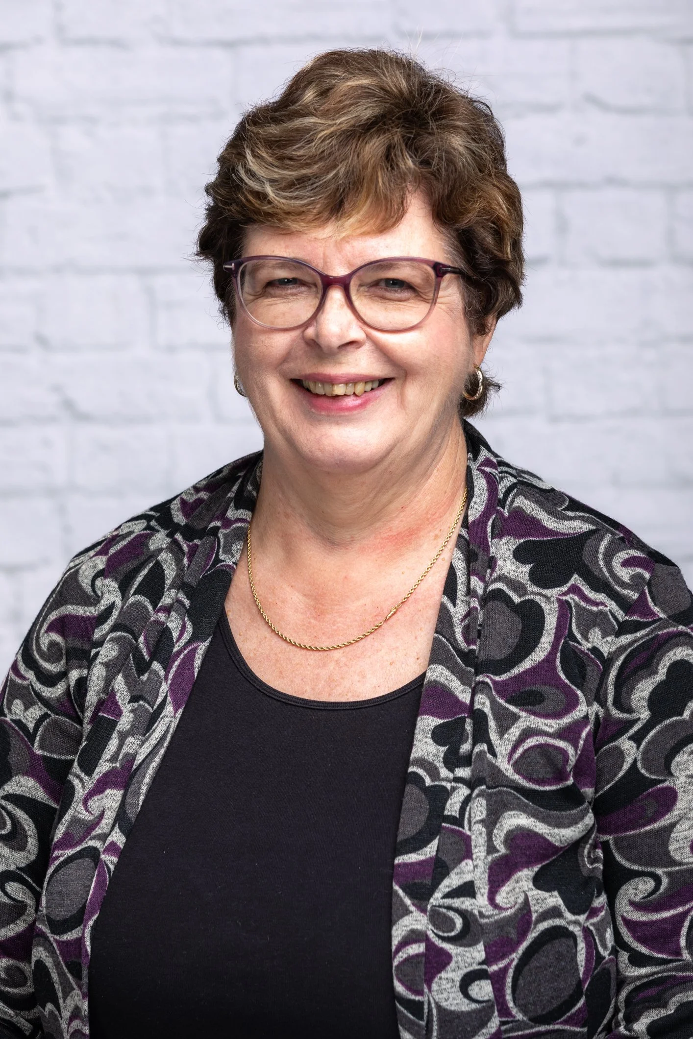 A middle-aged woman with short brown hair wearing glasses, a black top, a patterned blazer with purple, black, and gray designs, a gold necklace, and earrings, standing against a white brick wall background.