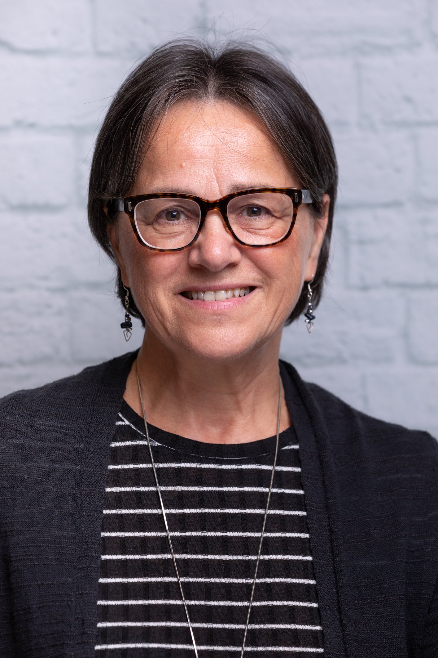 A close-up portrait of a middle-aged woman with short dark hair, wearing glasses, earrings, a black cardigan, and a black and white striped shirt, standing against a light gray brick wall.