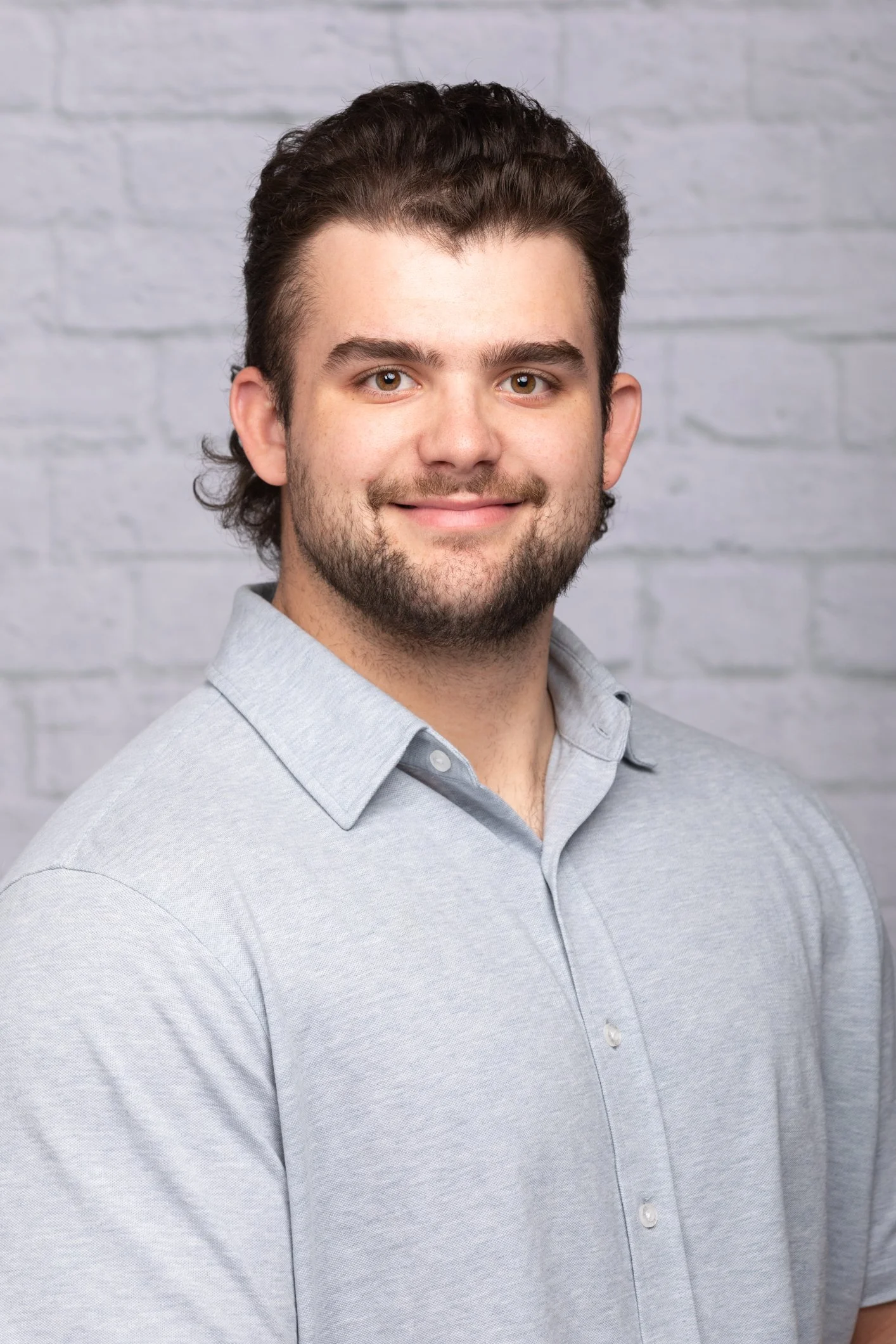 A smiling man with dark, wavy hair and a beard, wearing a light gray button-up shirt, standing in front of a light brick wall.