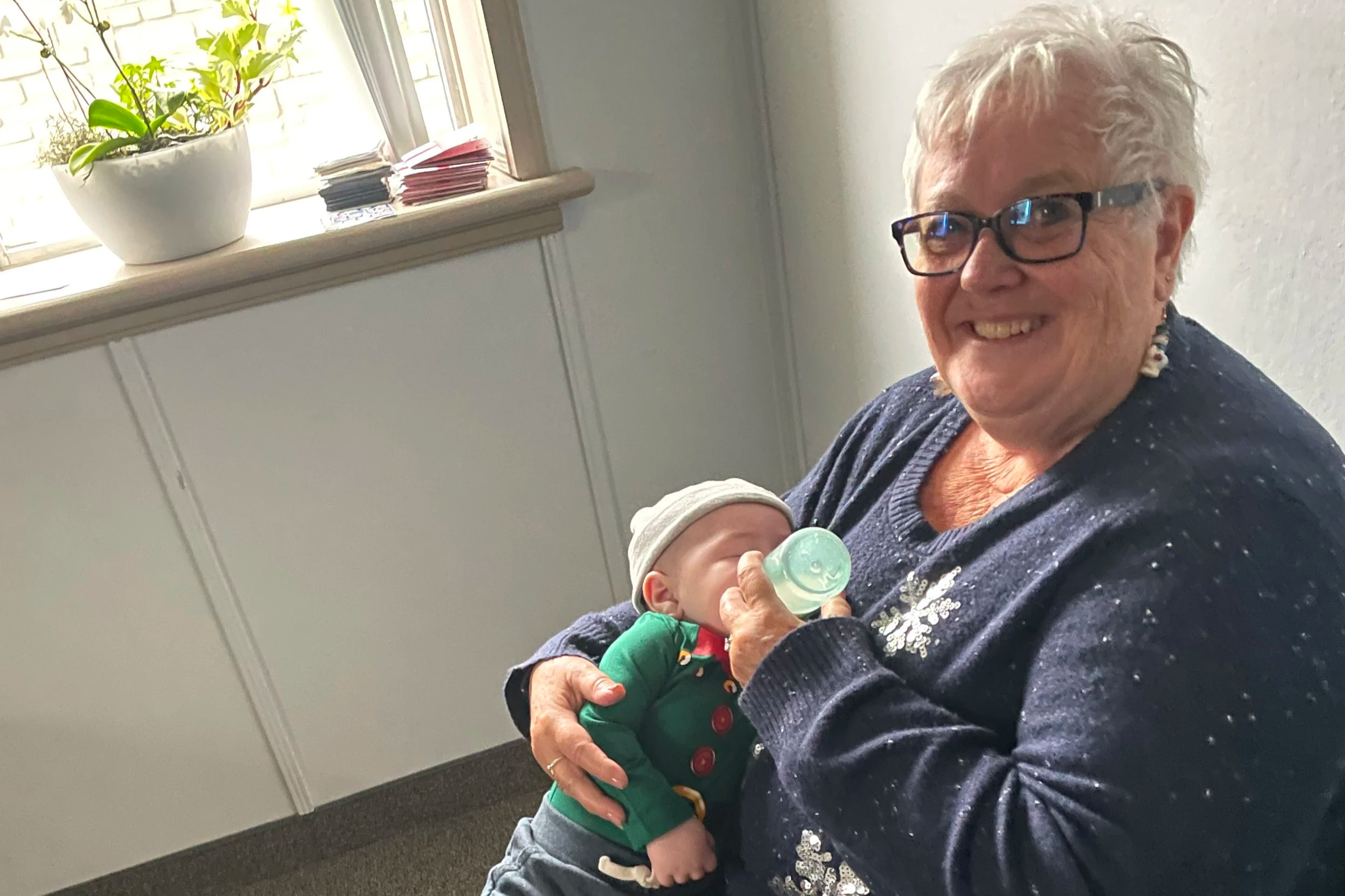 An elderly woman with white hair, glasses, and earrings smiles while holding a baby. The baby, dressed in green with a gray cap, is drinking from a baby bottle. They are indoors near a window with a potted plant and some stacked papers or cards on the windowsill.