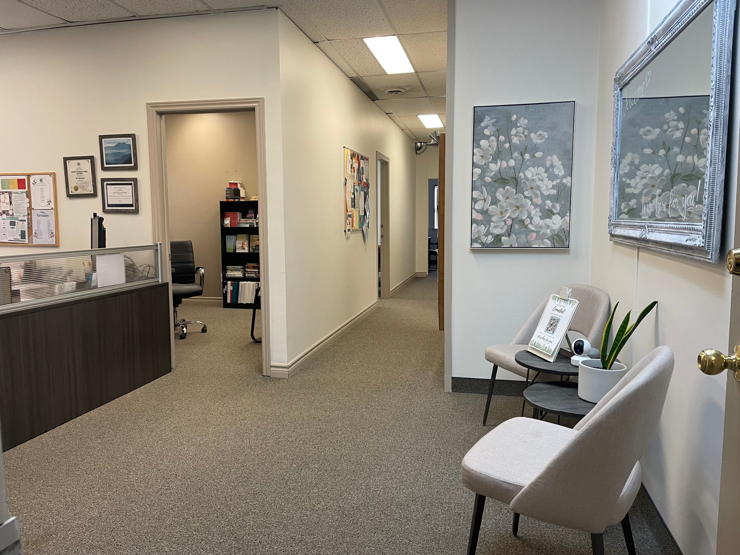 An office waiting area with two beige chairs, a small table with a plant, framed artworks on the wall, and a corridor leading to other rooms.