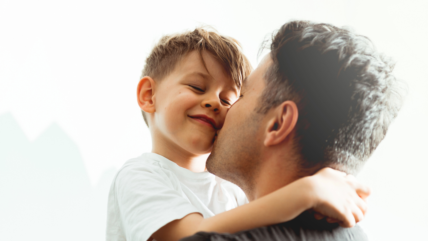 A young boy with light brown hair smiling and hugging an adult man, possibly his father, who has short, curly gray hair.