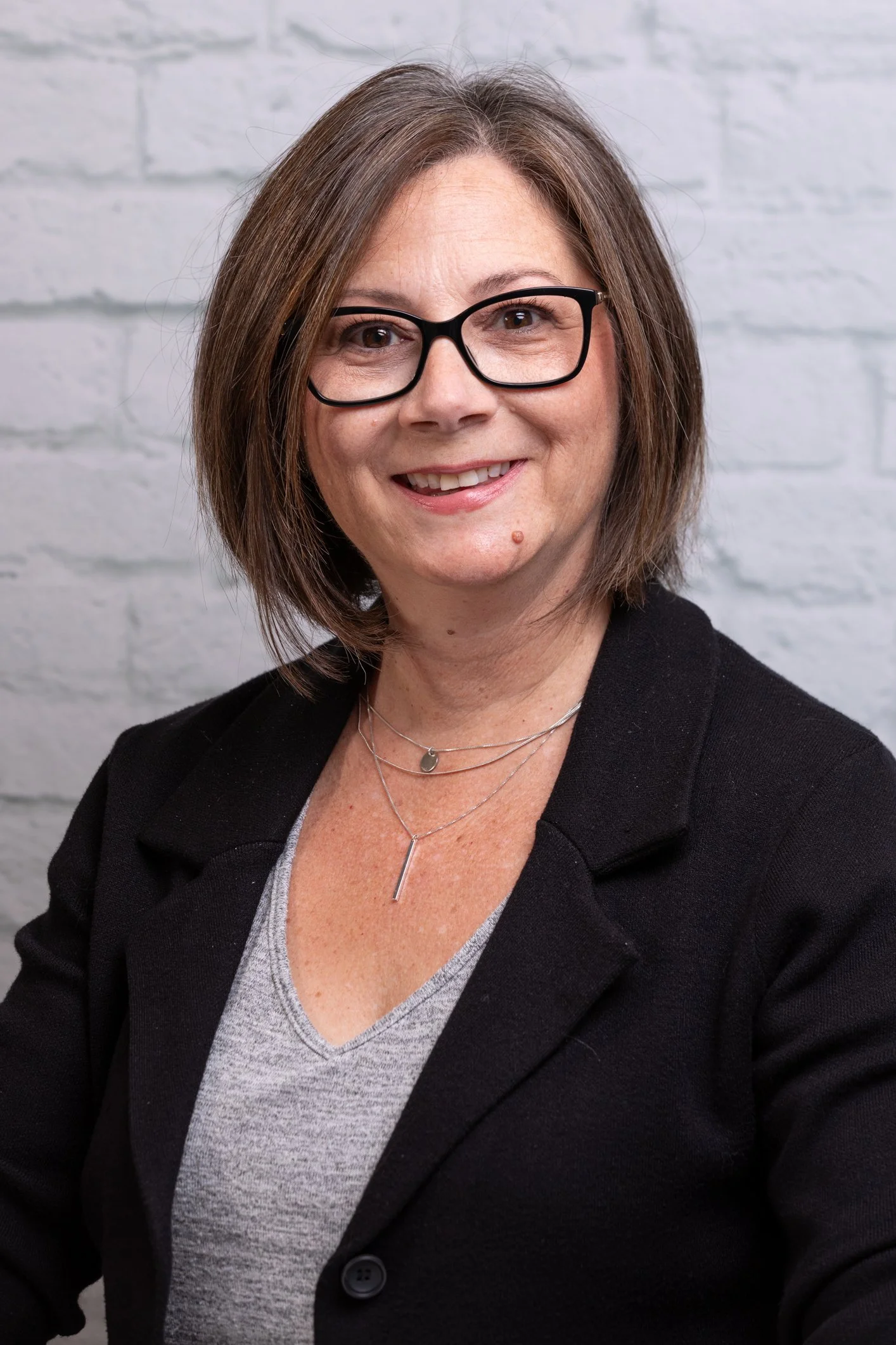 Portrait of a woman with glasses smiling in front of a white brick wall, wearing a black blazer and layered necklaces.