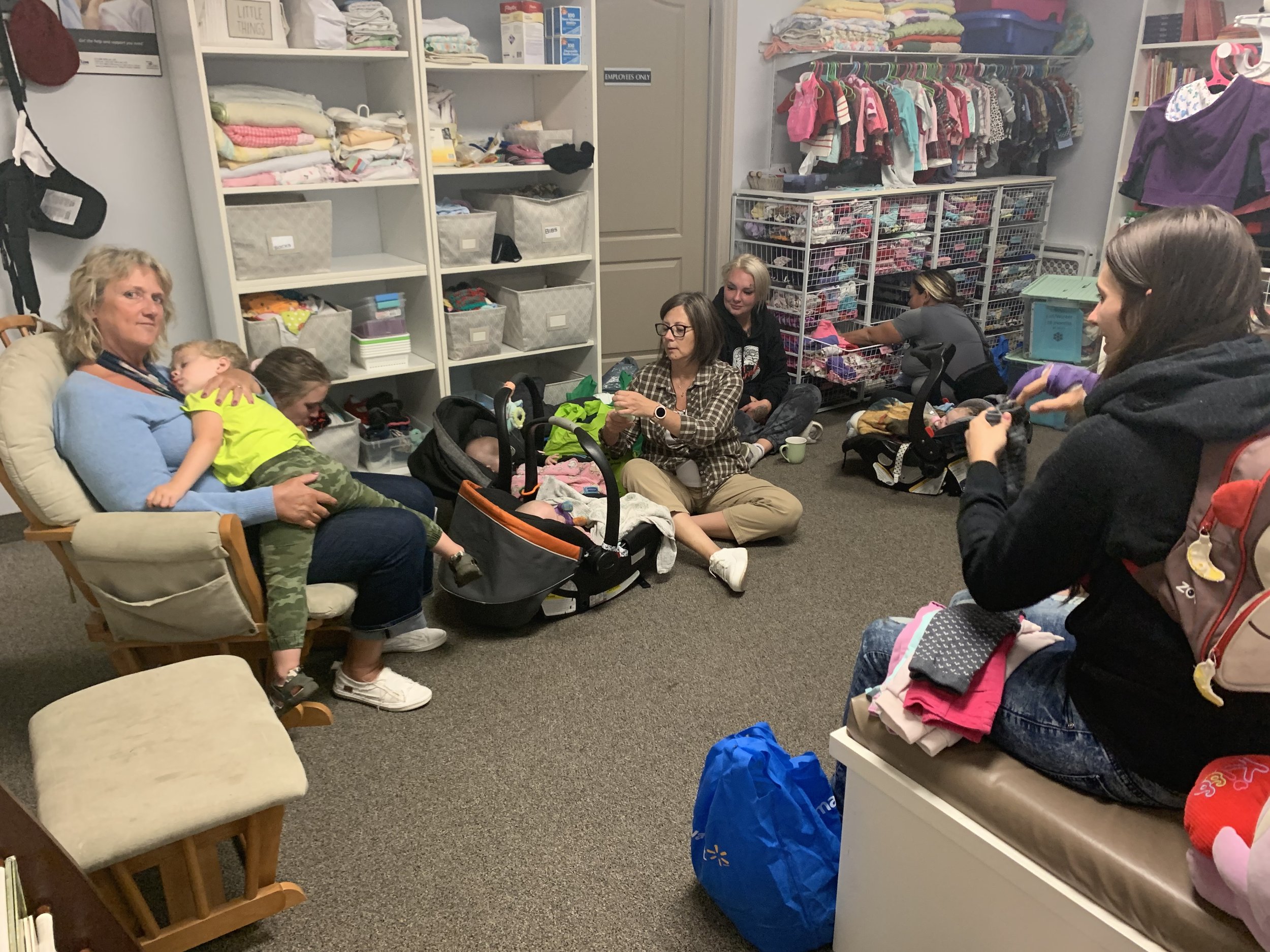 A group of women and children inside a room filled with shelves of toys and clothes. One woman is sitting on a chair holding a young boy, possibly sleeping, on her lap. Others are sitting on the floor, organizing or sorting items, with some children in car seats.
