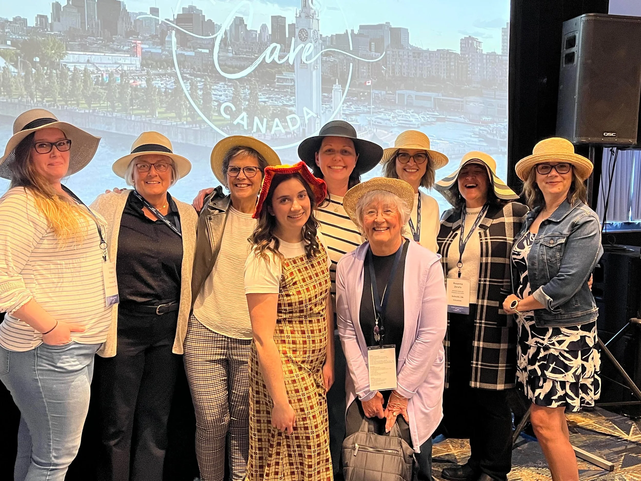 A group of ten women wearing various styles of hats, standing together at an indoor event with a large screen displaying a cityscape and the words 'Care Canada' in the background.