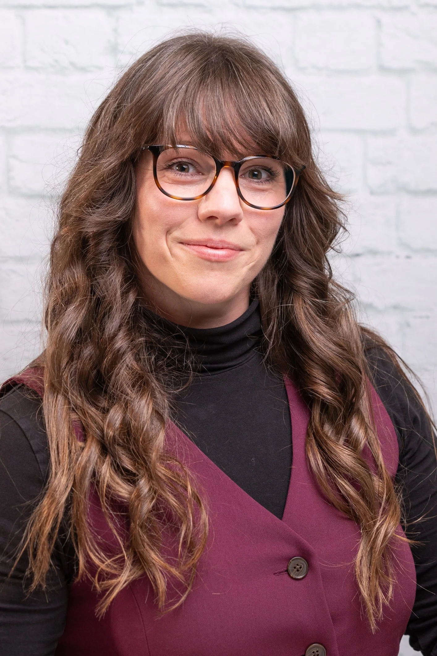 A woman with long, wavy brown hair and glasses, smiling, wearing a black turtleneck under a maroon button-up vest, standing against a white brick wall.