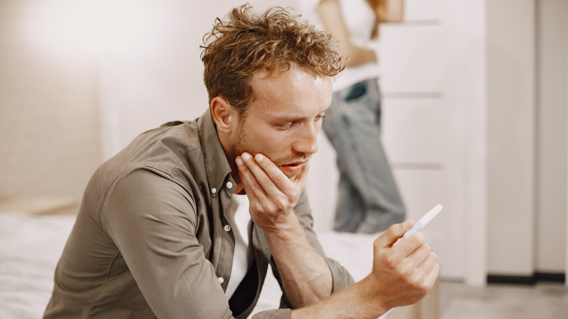 A man sitting on a bed looking at a pregnancy test with a concerned expression, in a bedroom.